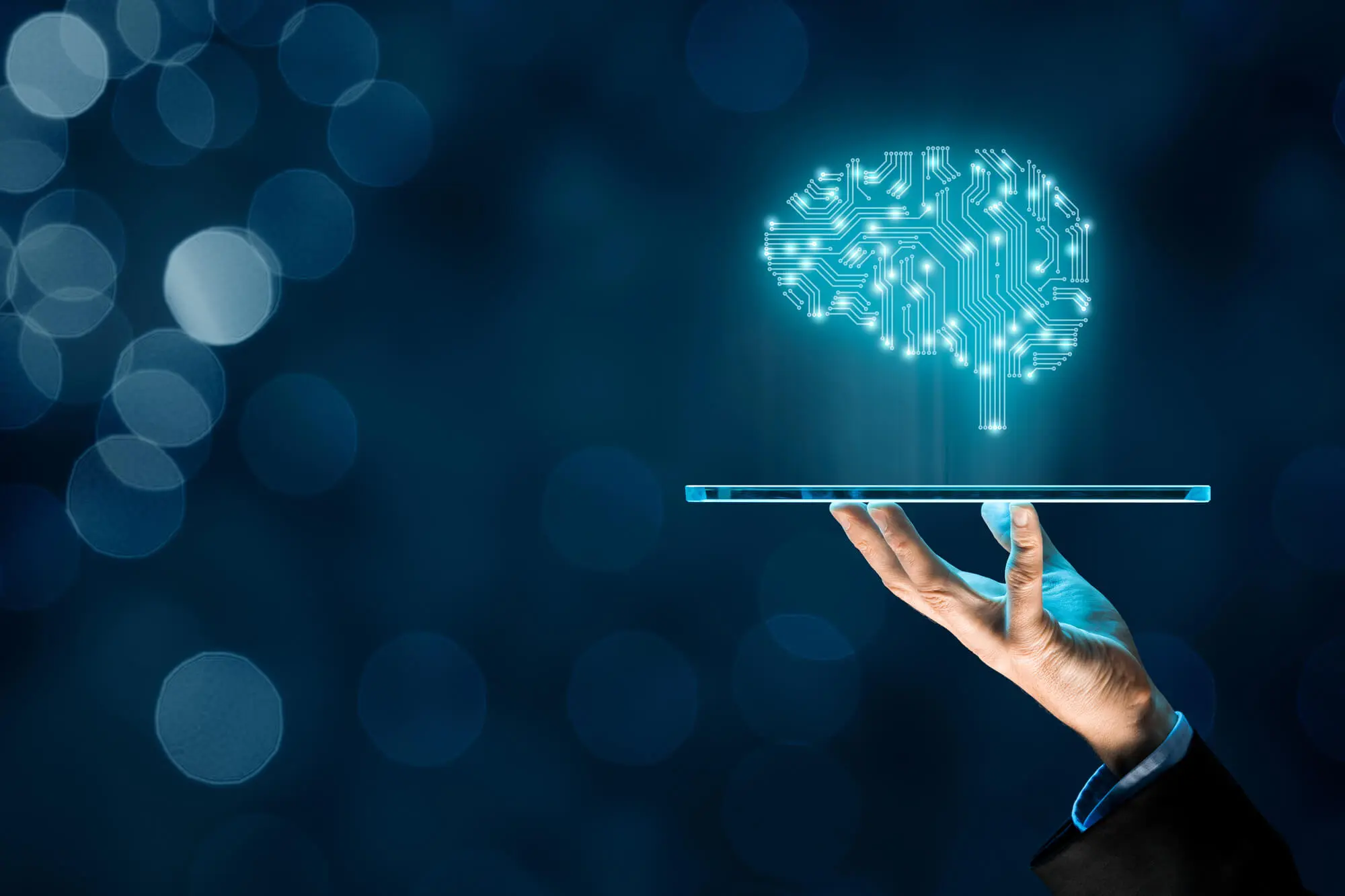 Hand holding a transparent tablet projecting a glowing digital brain with circuitry on a blue bokeh background