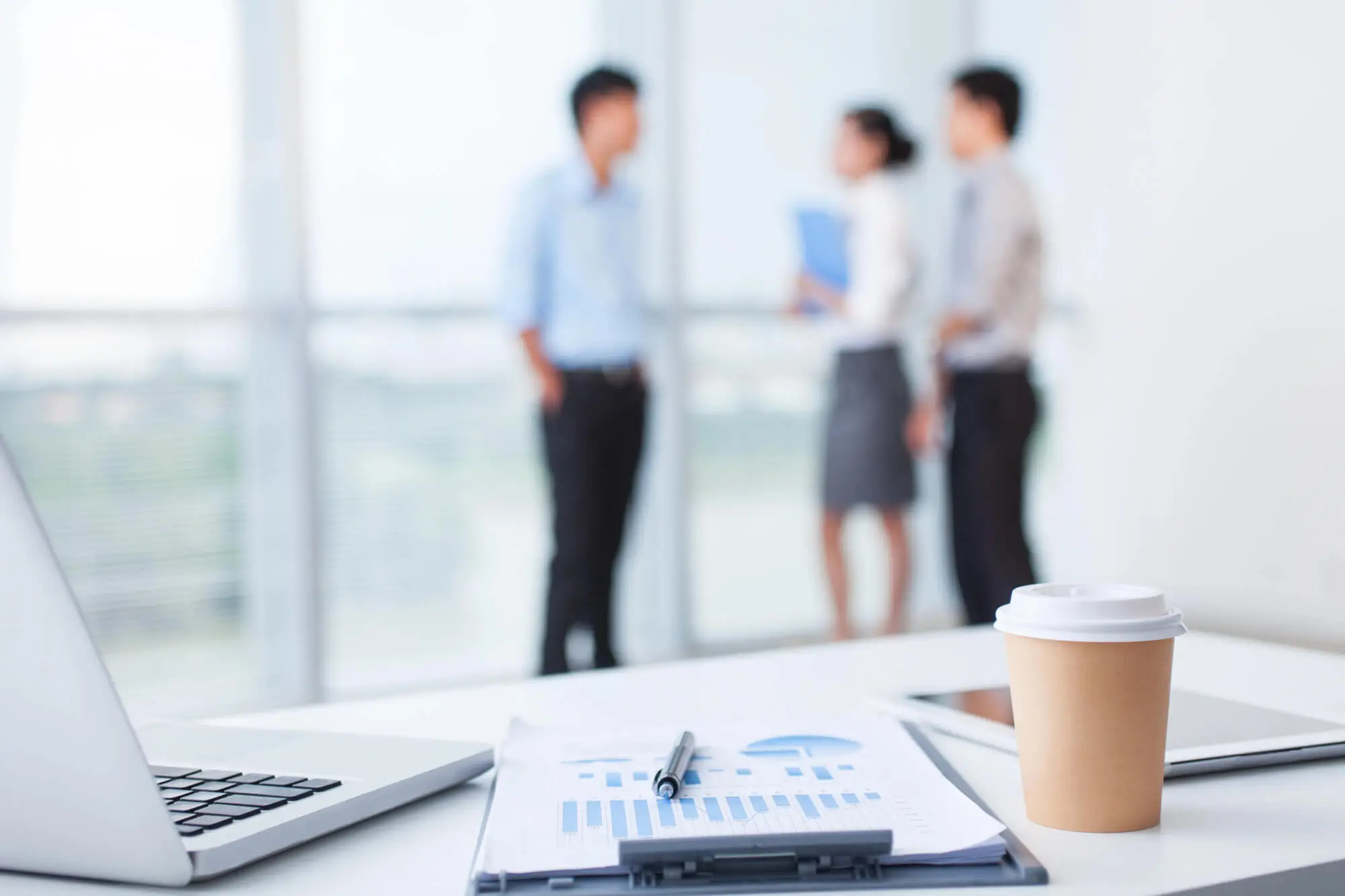 Office desk with laptop, documents, pen, coffee cup, and tablet; three blurred coworkers stand in the background near large windows