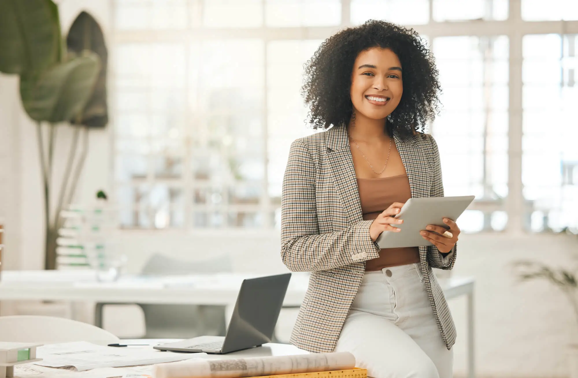 Smiling woman in a plaid blazer holds a tablet, leaning on a desk with a laptop. Bright, modern office with large windows and a relaxed atmosphere