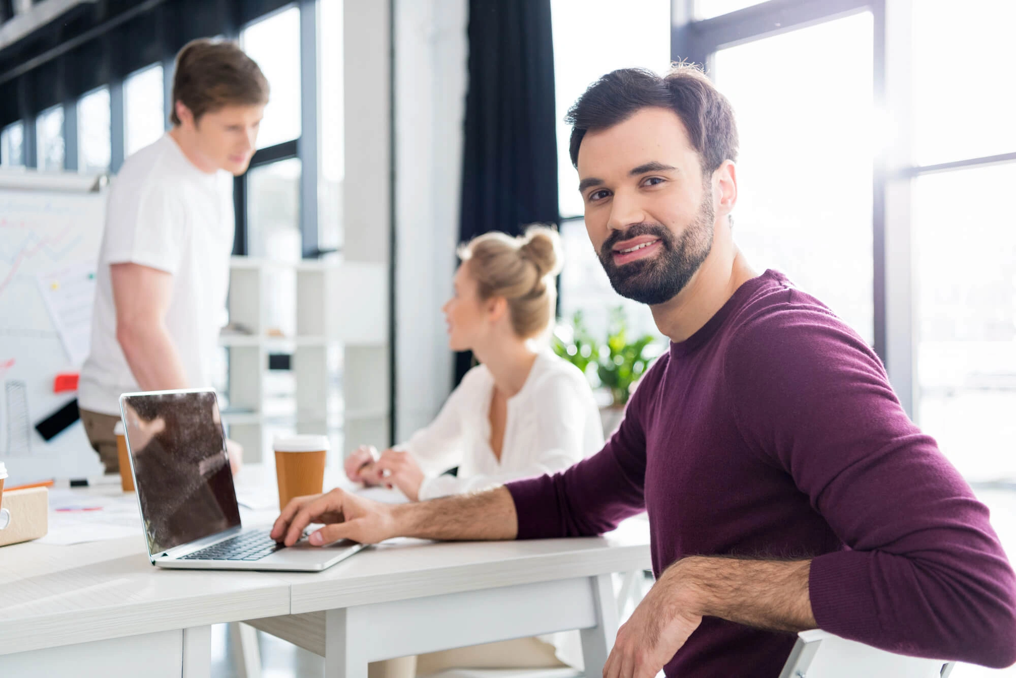 Man in a maroon sweater smiling at a desk with a laptop. Two colleagues in discussion in the background. Bright, modern office setting conveys teamwork