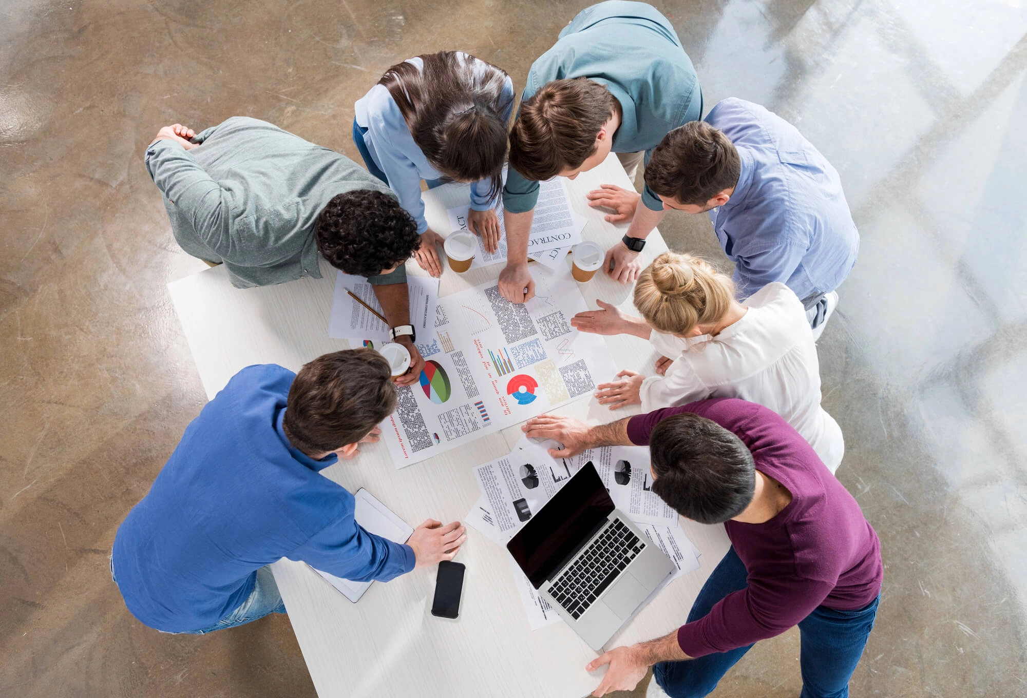 Overhead view of a diverse group collaborating around a table with charts, graphs, and a laptop. The scene conveys teamwork and focused discussion