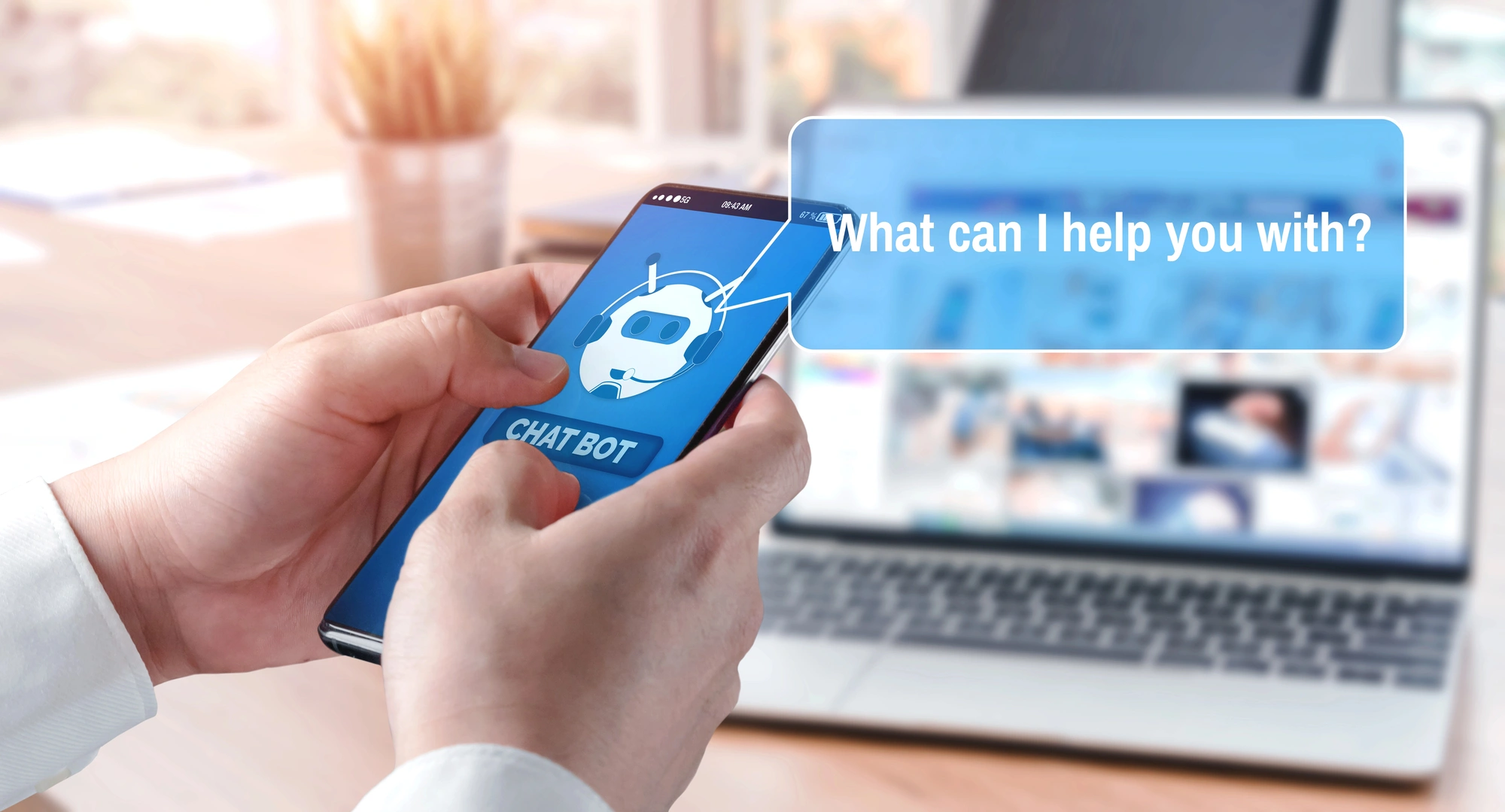 A person holds a smartphone displaying a chatbot image. A chat bubble asks, "What can I help you with?" A laptop is blurred in the background