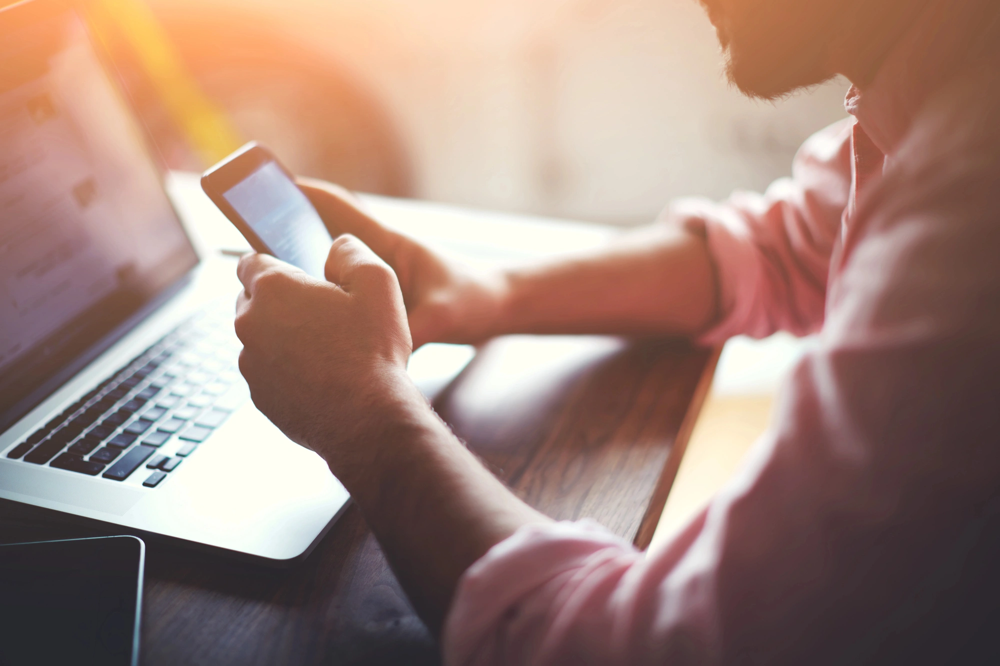 A person in a pink shirt uses a smartphone at a wooden desk with an open laptop. Warm sunlight filters through, creating a focused, serene atmosphere