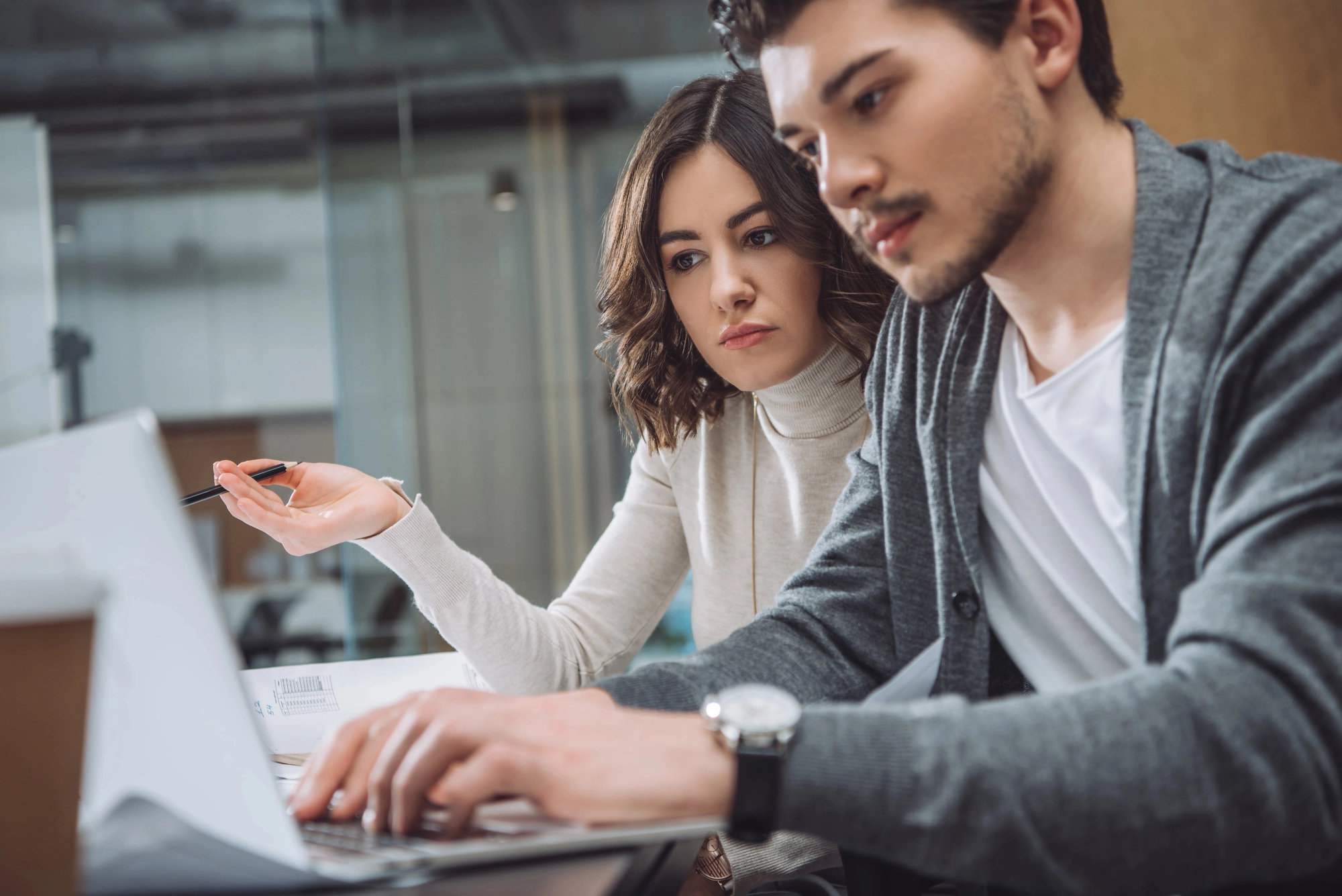 Two people working together on a laptop, one typing and the other gesturing with a pen in an office setting
