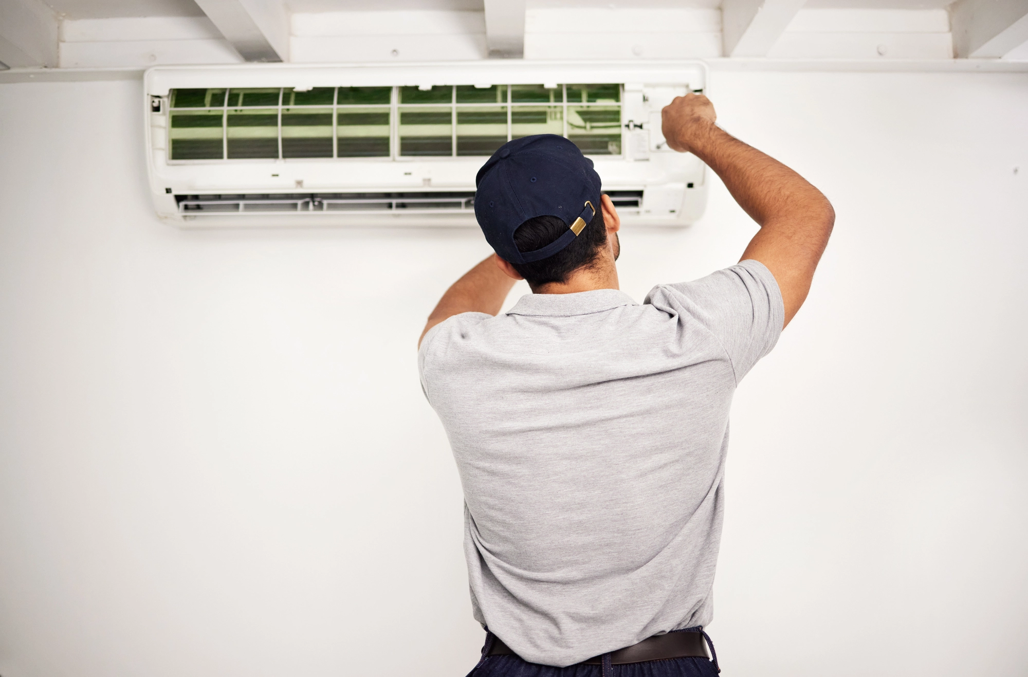Technician in a cap and grey shirt repairing or inspecting a wall-mounted air conditioner unit