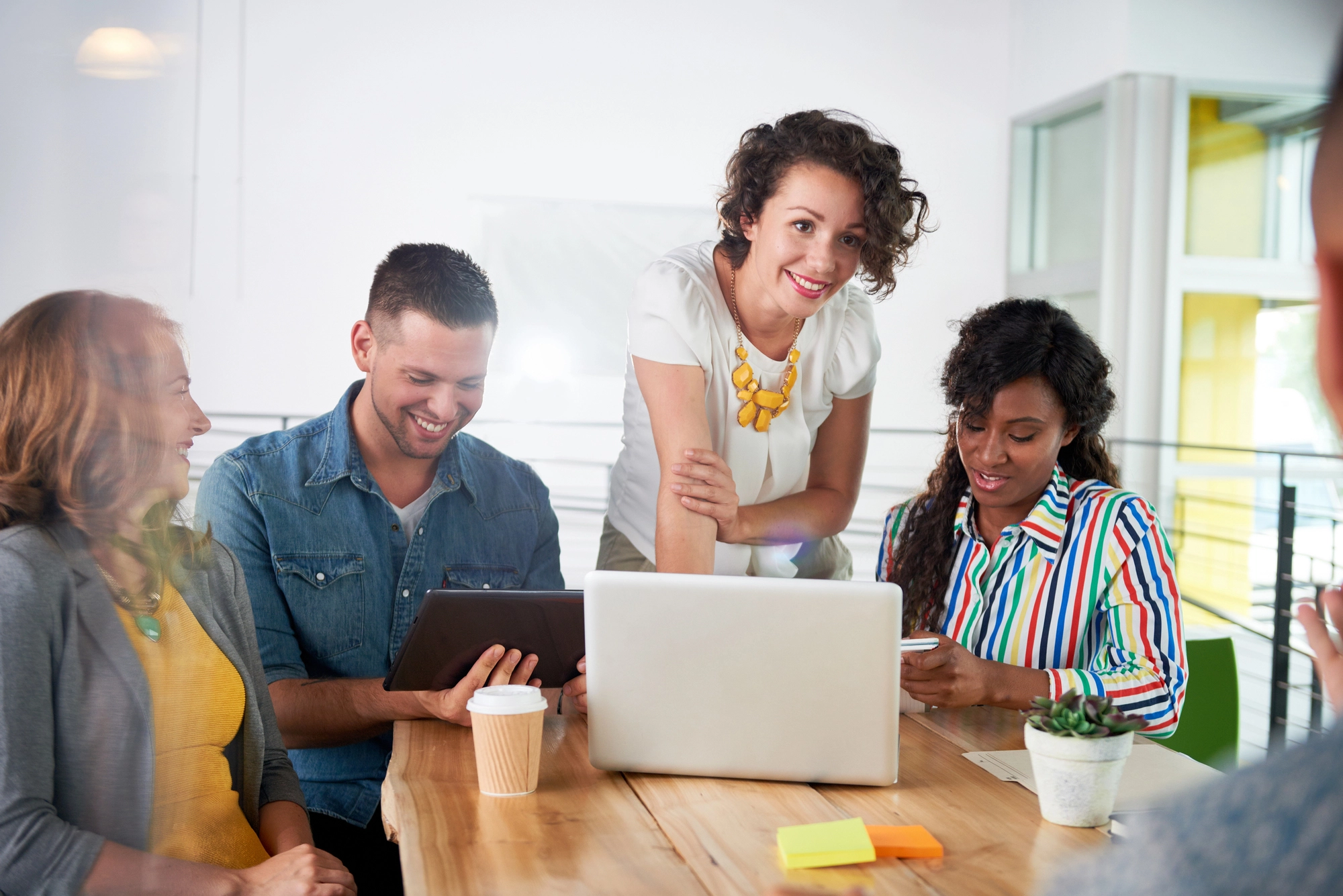 Group of diverse colleagues collaborating around a table with laptop, tablet, coffee cup, and plant in a modern office setting