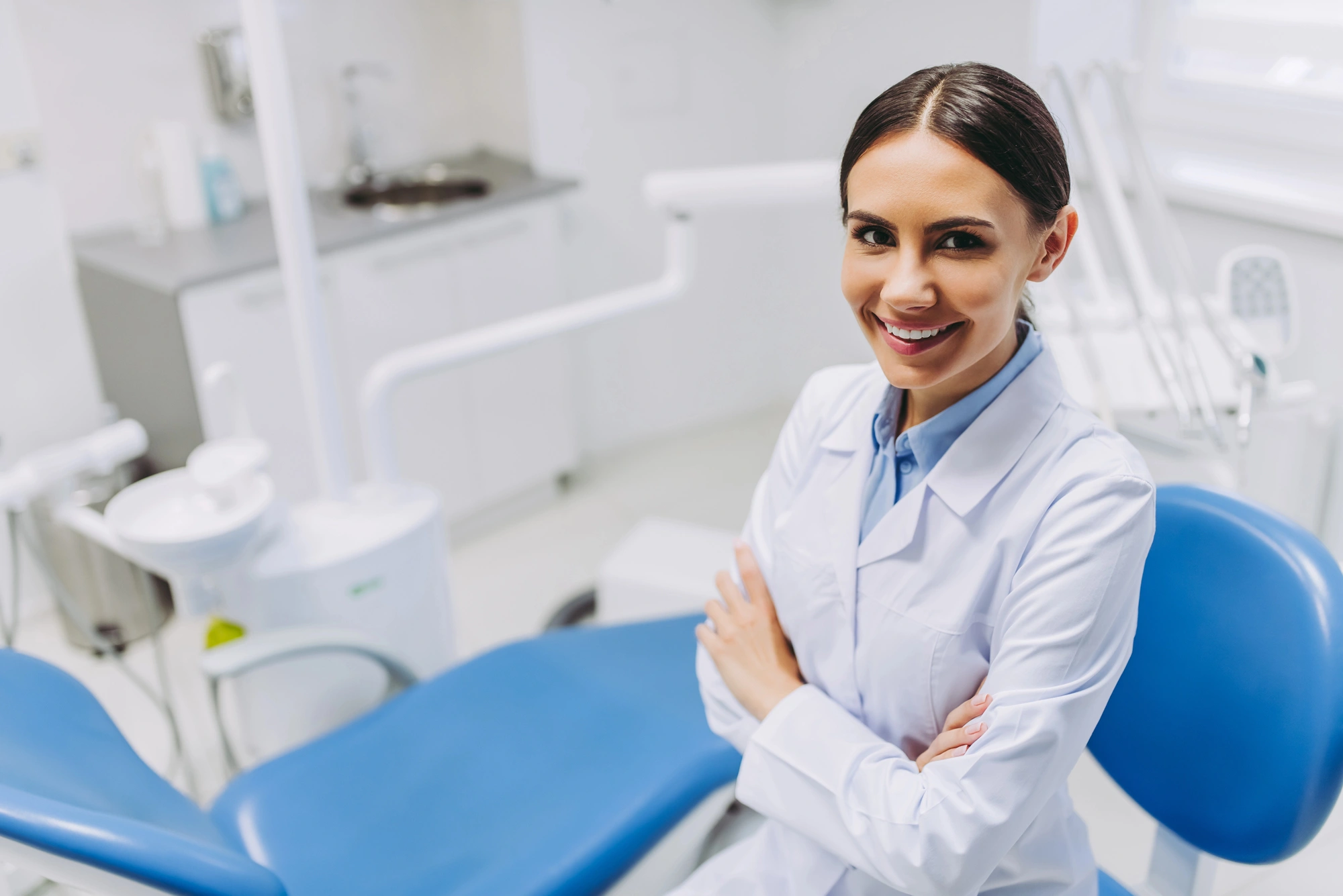 Female dentist in white coat sitting with arms crossed next to a blue dental chair in a bright clinic room