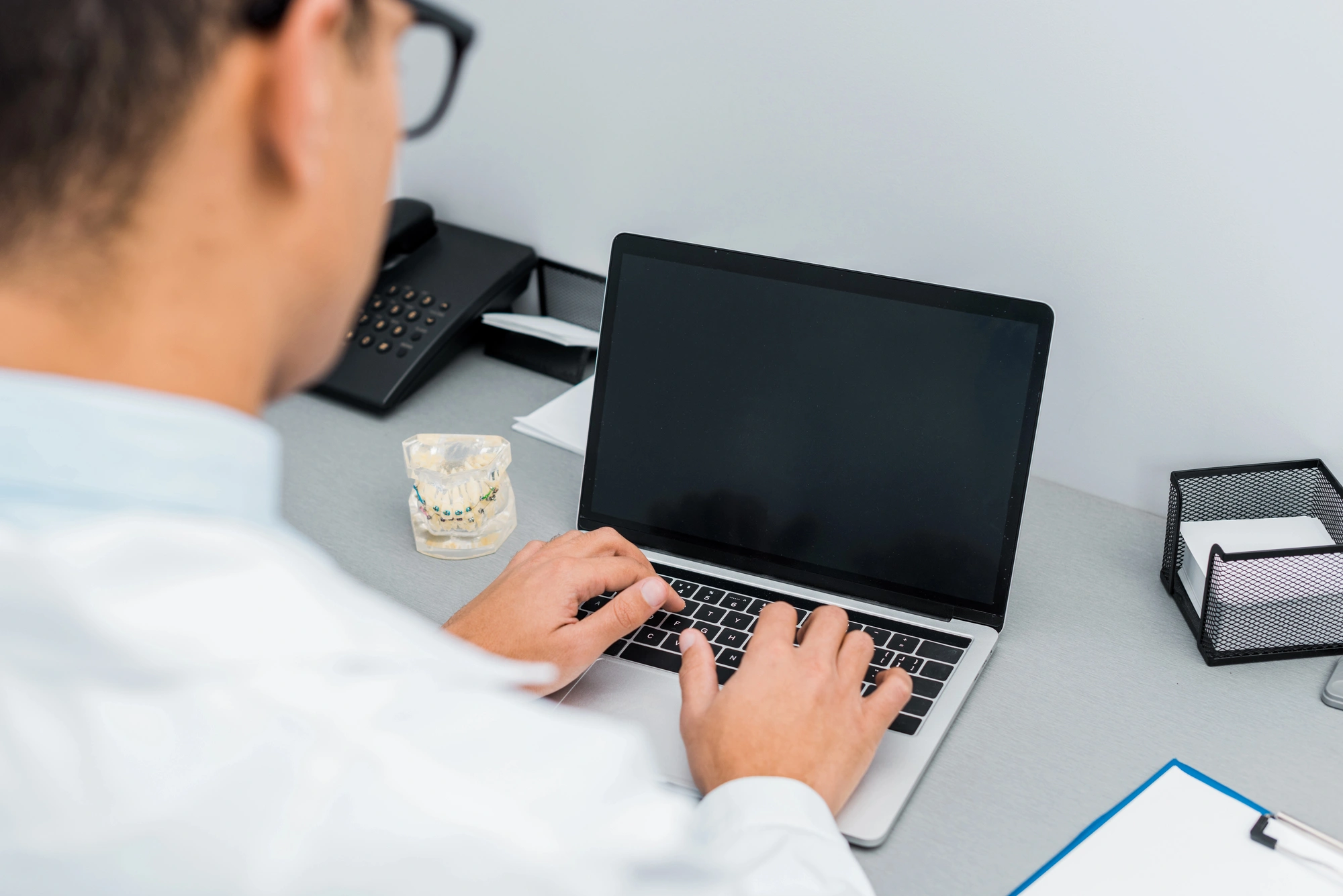 Man in white shirt typing on a laptop at a desk with dental model, phone, and office supplies