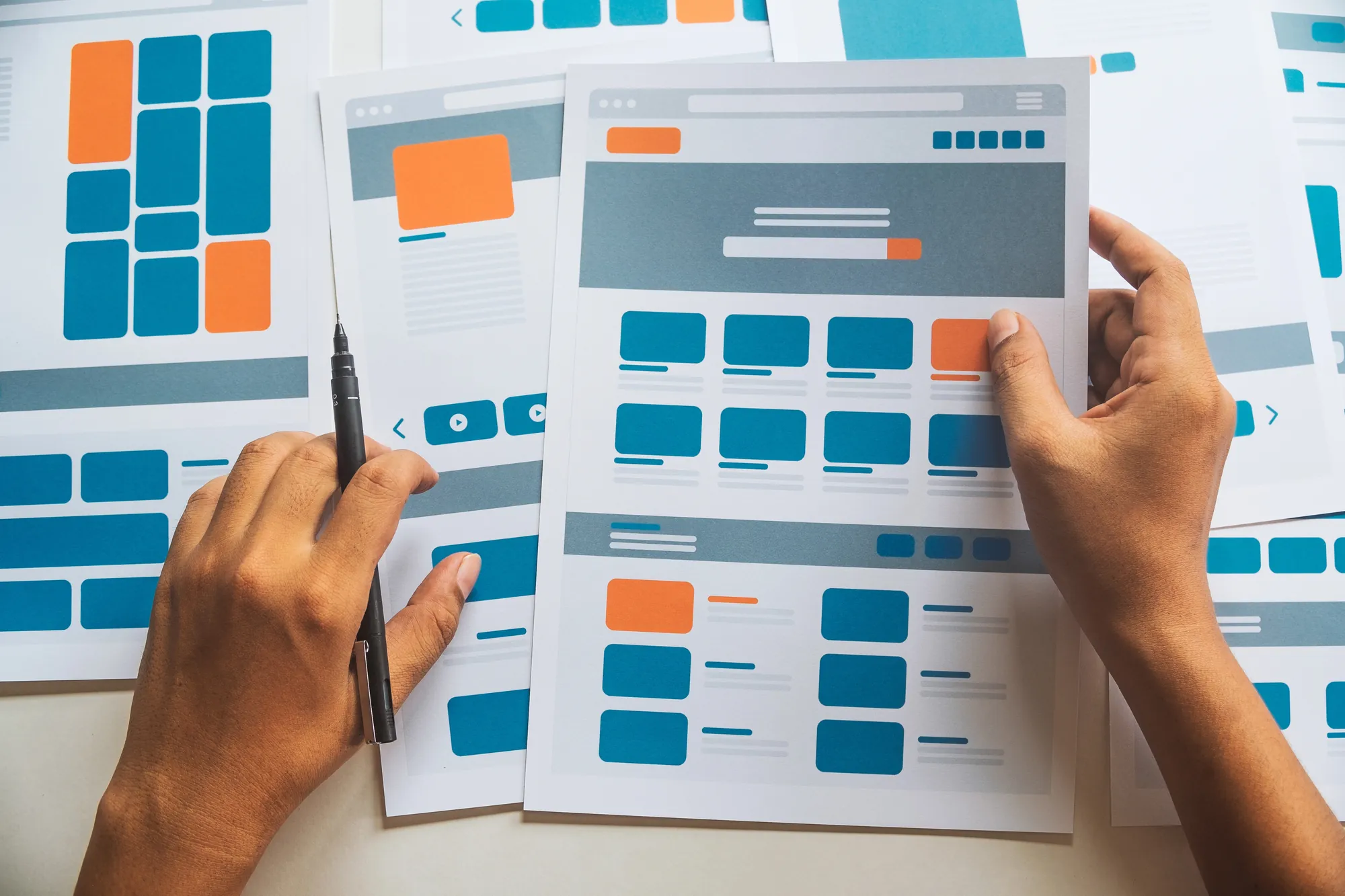 Close-up of hands reviewing printed website wireframe layouts with orange and blue design blocks and a marker pen