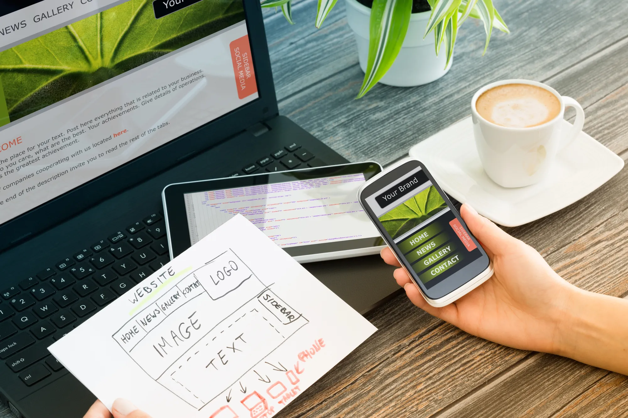 Hand holding a smartphone showing a mobile website preview next to a laptop, tablet, hand-drawn wireframe sketch, and coffee on a wooden desk