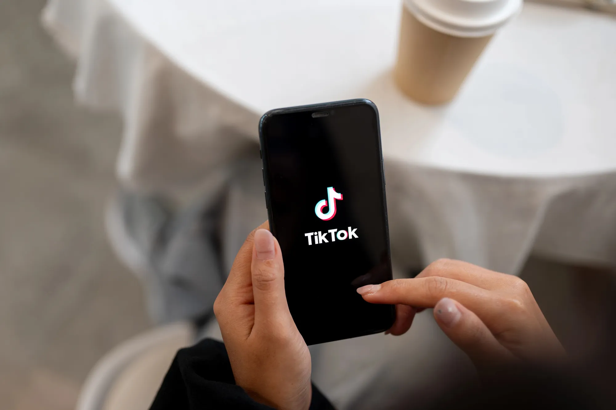 Hand holding a smartphone displaying the TikTok logo splash screen at a cafe table with a coffee cup nearby