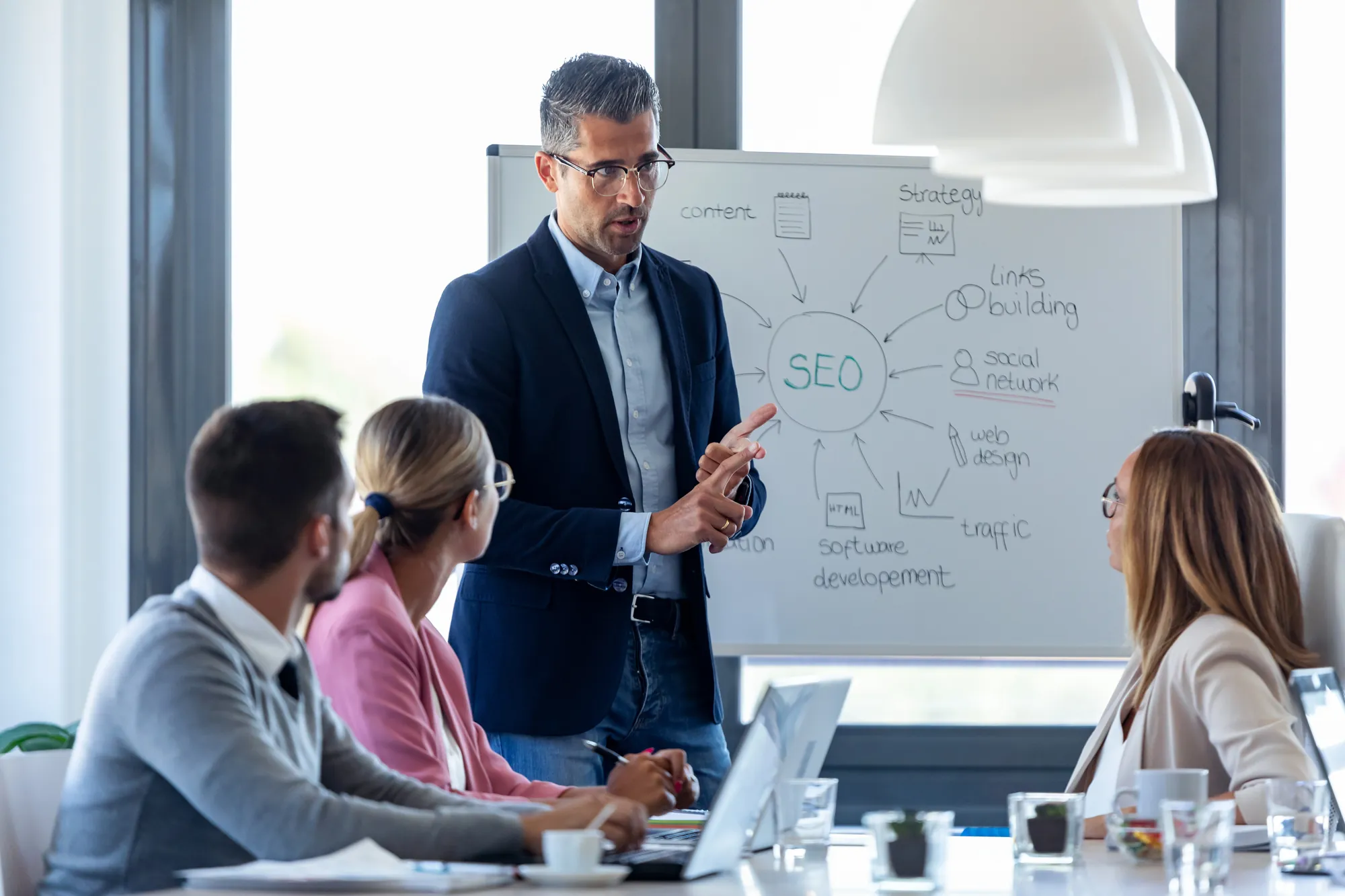 Business consultant presenting an SEO strategy diagram on a whiteboard to colleagues in a meeting room