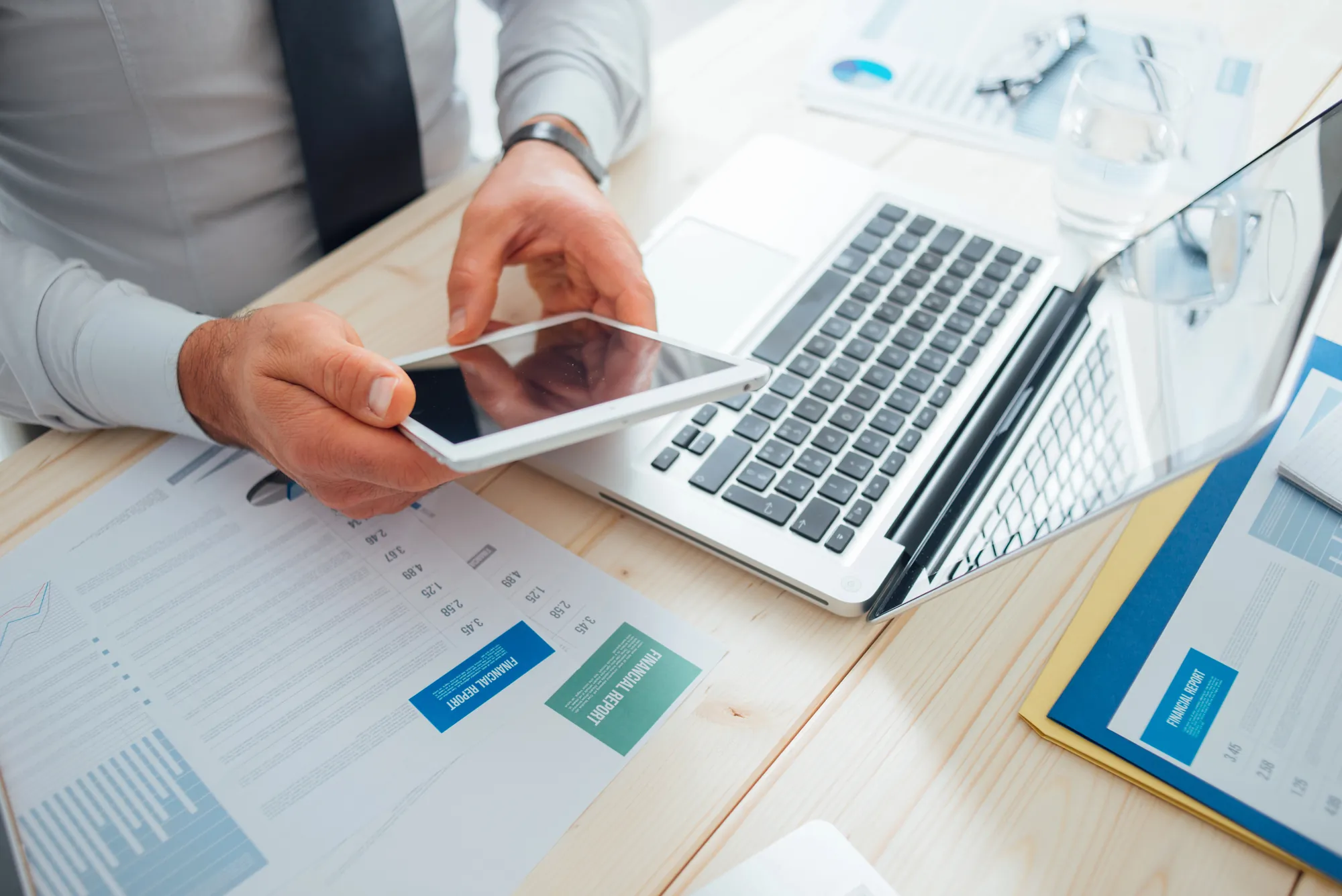 Close-up of a businessman holding a tablet next to a laptop and financial report documents on a desk