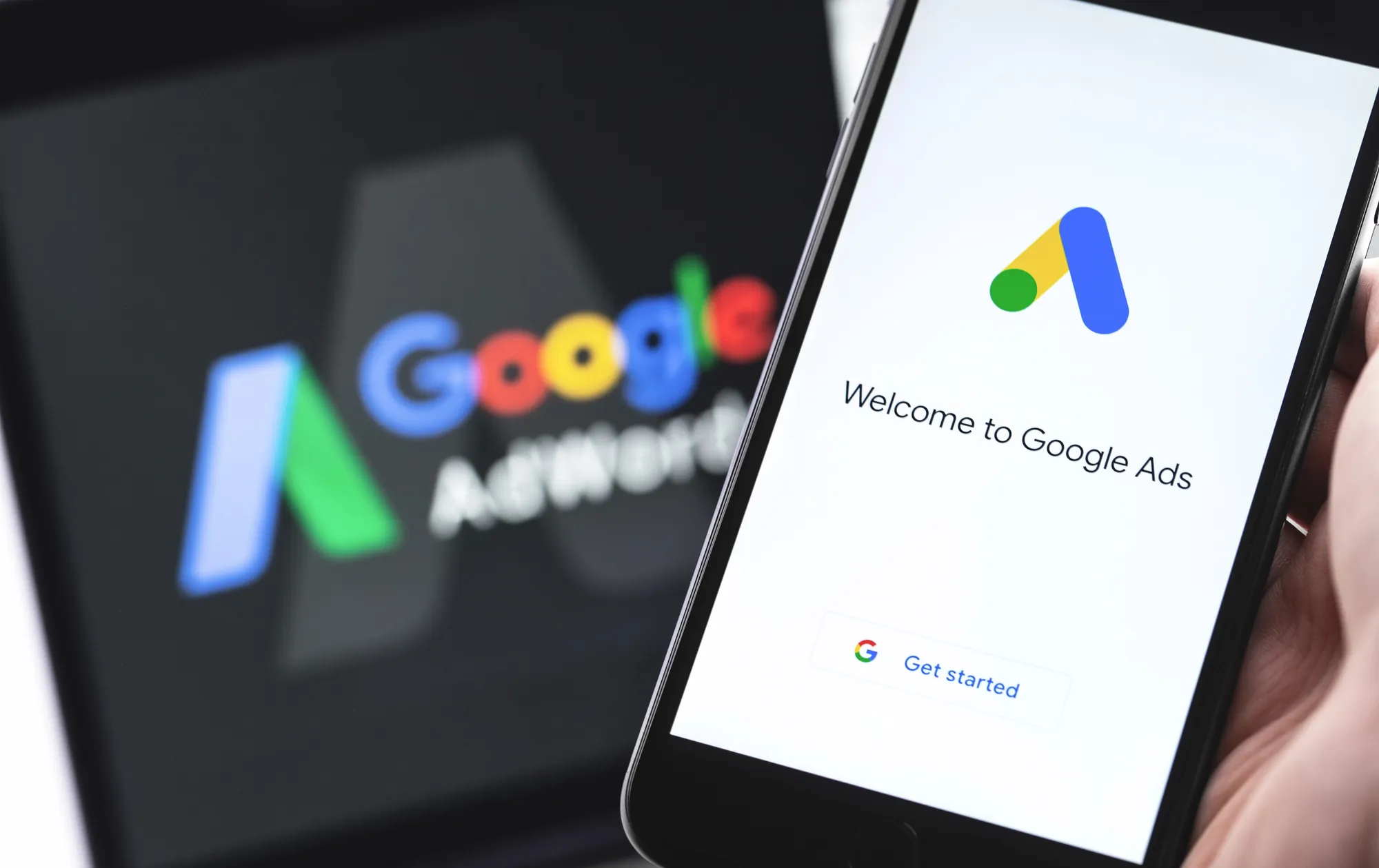 Hand holding a smartphone showing the Welcome to Google Ads screen with a blurred Google Ads logo in the background