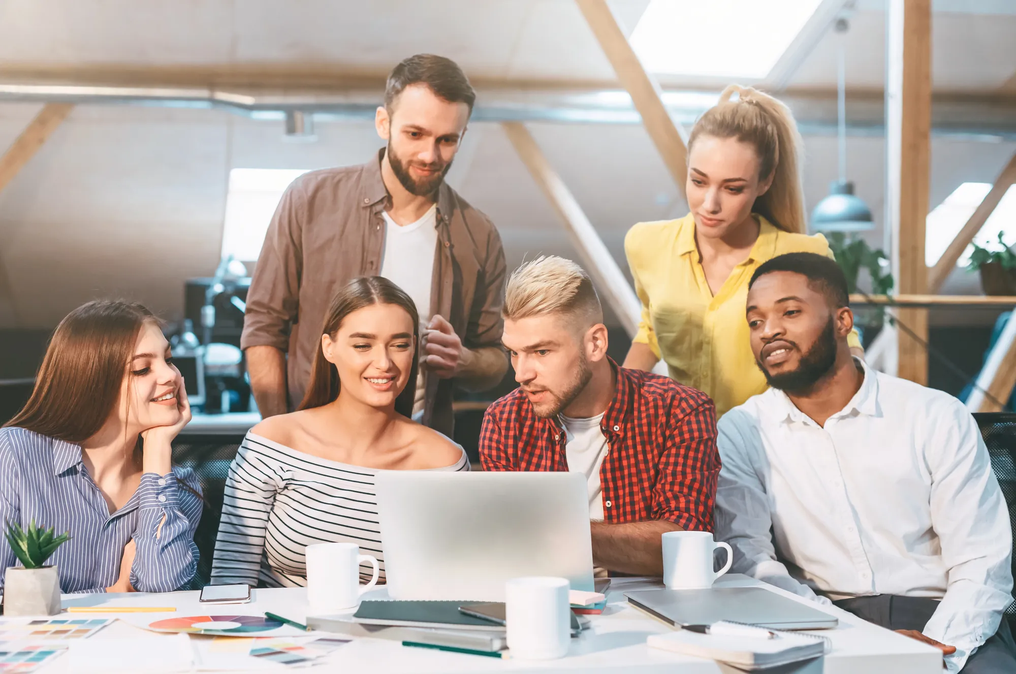 Group of six coworkers gathered around a laptop discussing a project in a bright office