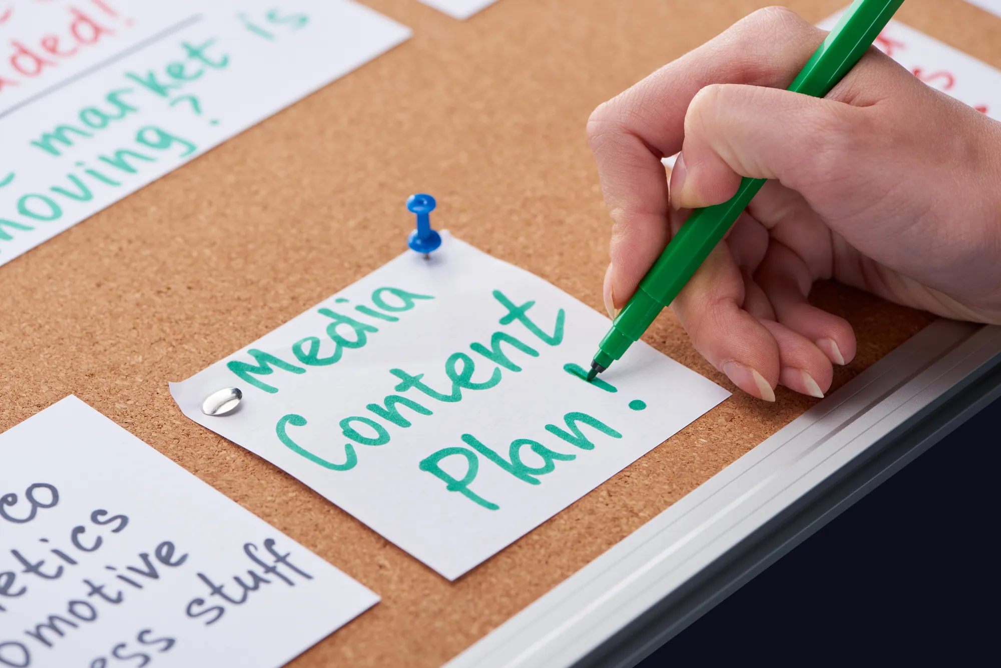 Close-up of a hand writing Media Content Plan on a sticky note pinned to a corkboard with other planning notes