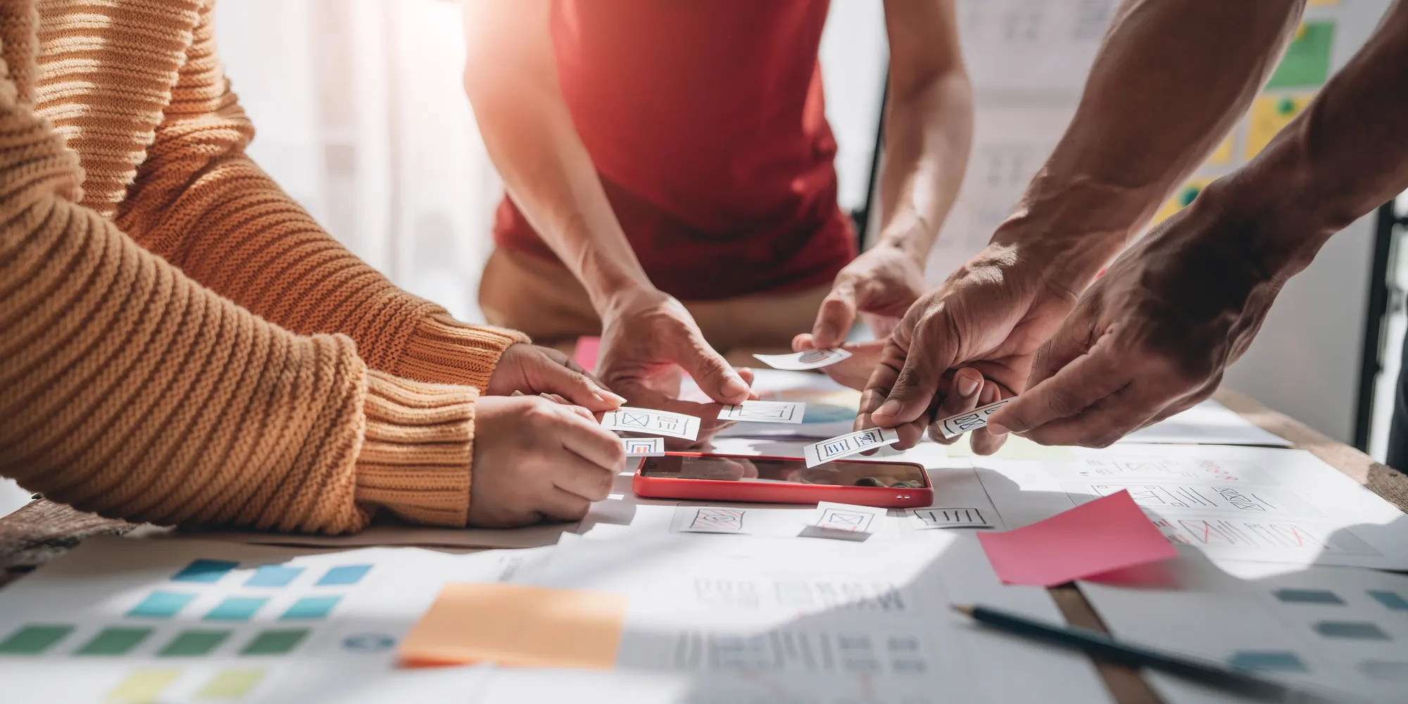 lose-up of hands arranging paper UX wireframe sketches around a smartphone on a desk covered in color swatches and sticky notes
