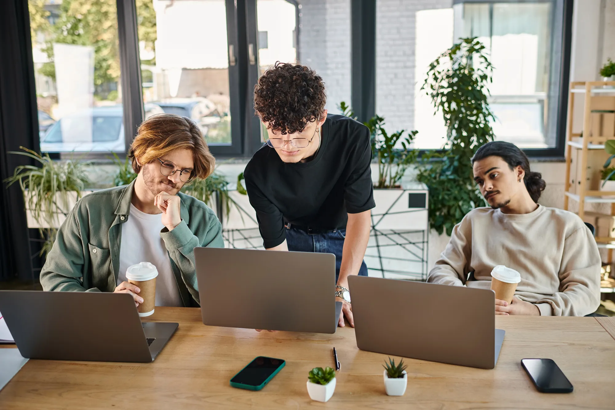 Three coworkers reviewing content on laptops together at a table in a bright office with coffee cups nearby