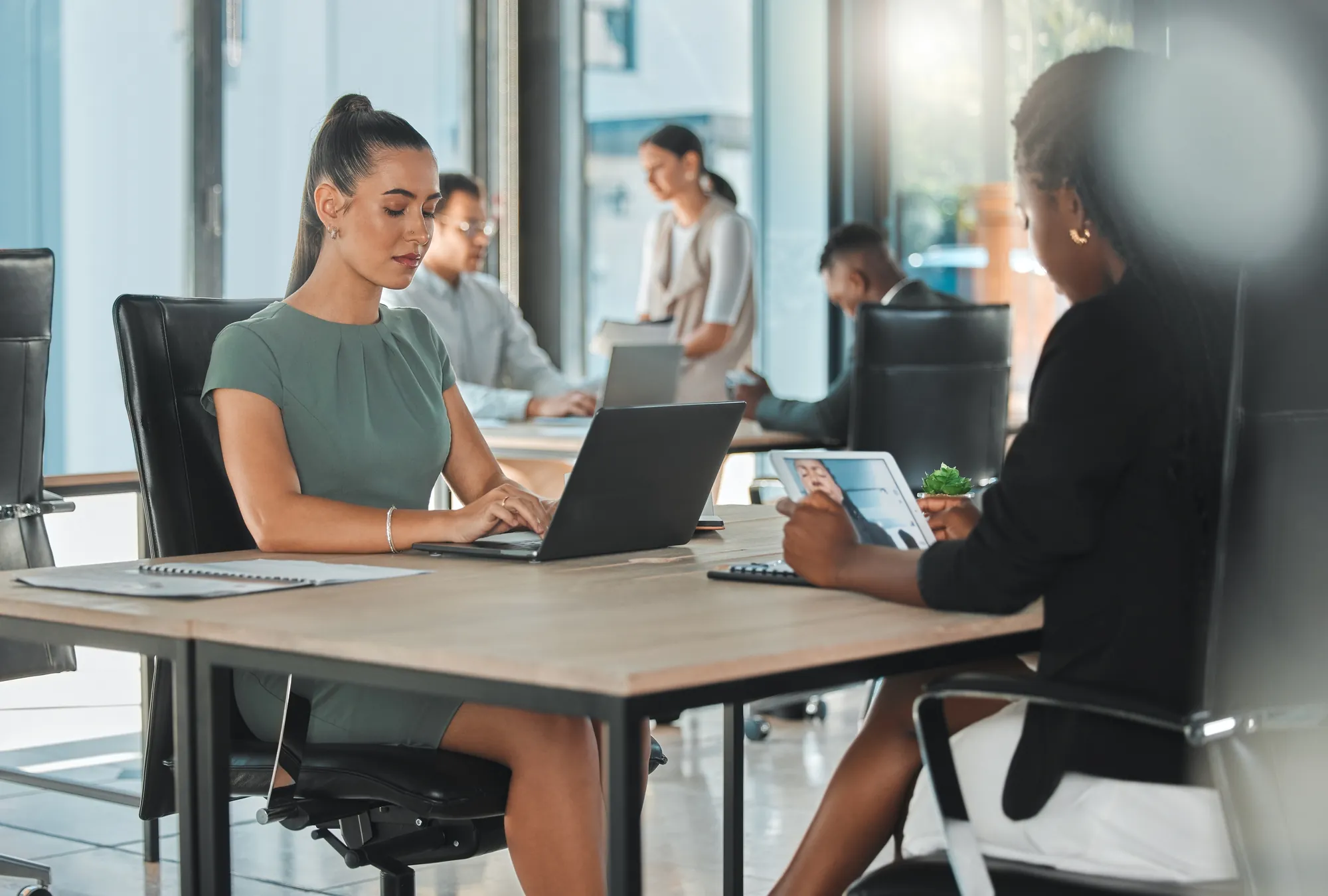 Two businesswomen working at desks with a laptop and tablet in a busy modern office