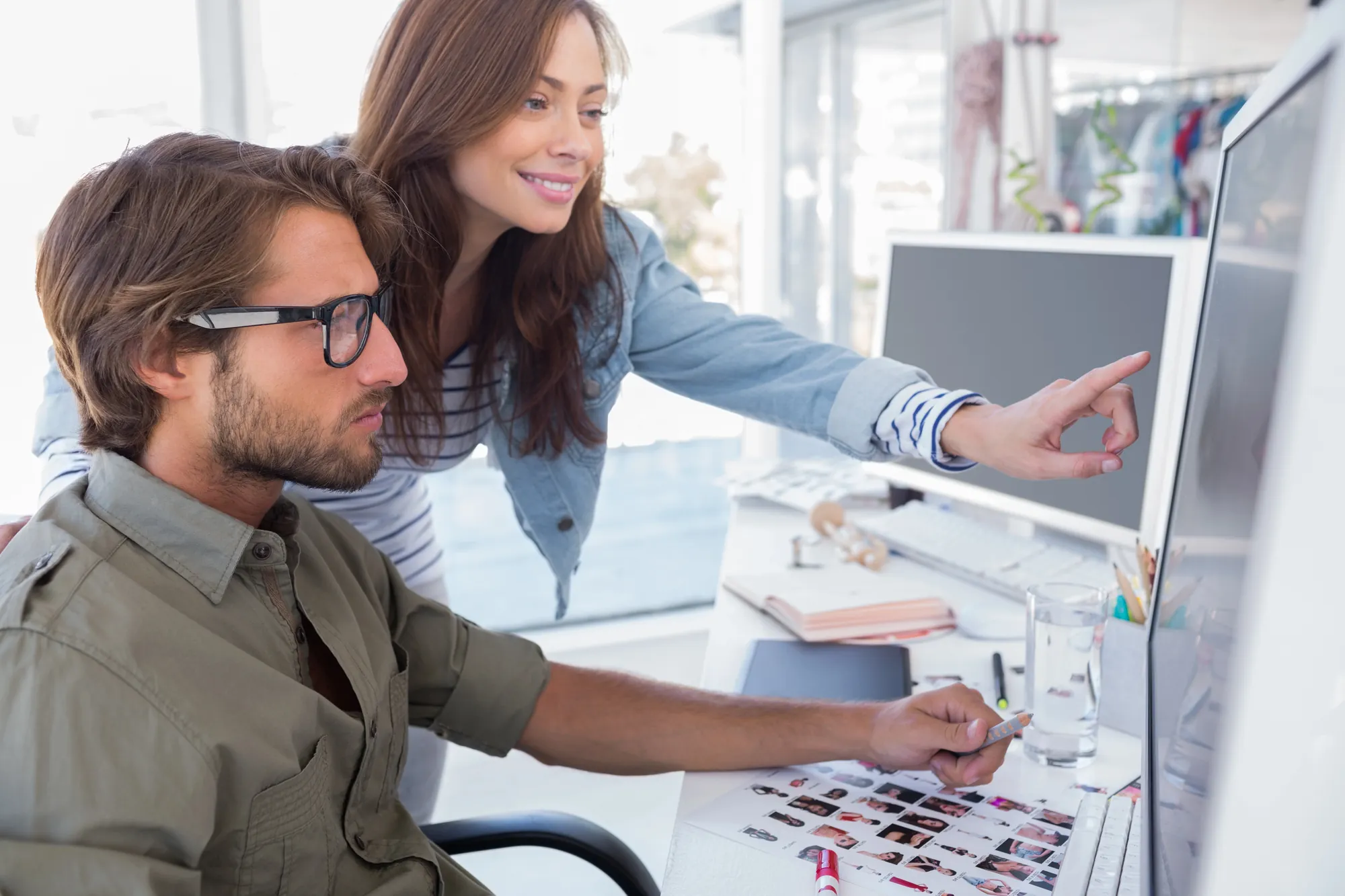Woman pointing at a computer monitor while a coworker reviews printed photo contact sheets at a desk