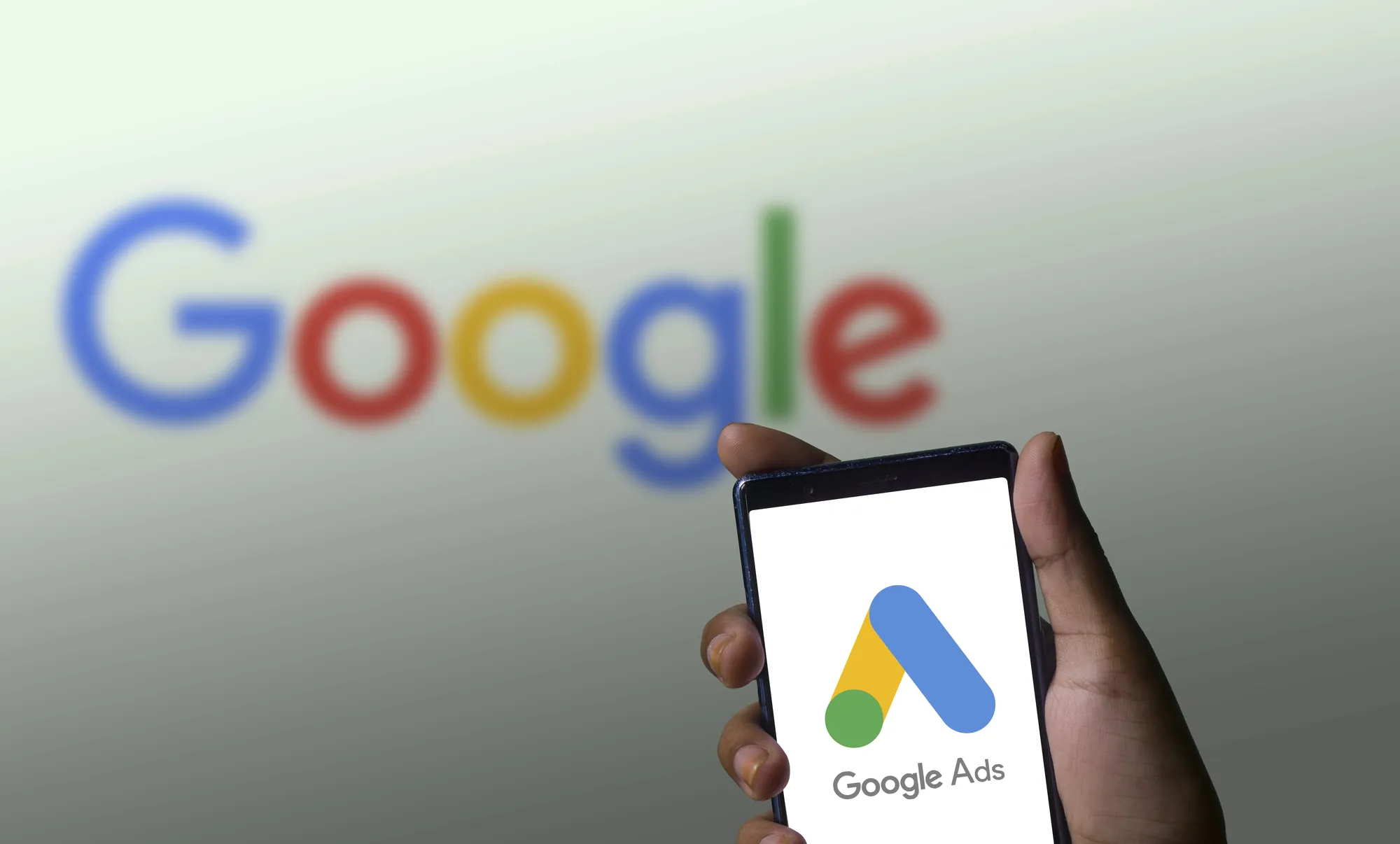 Hand holding a smartphone showing the Google Ads logo with the blurred Google logo displayed on a screen behind it