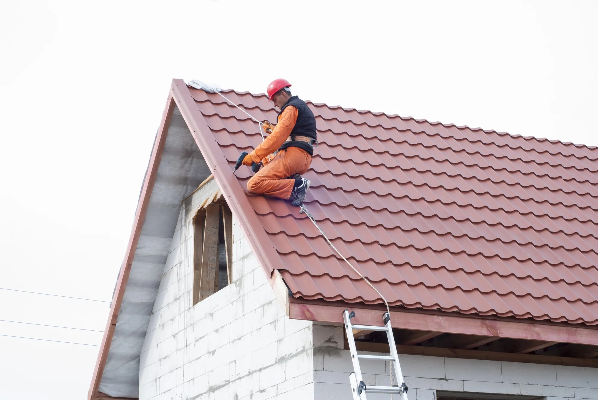 Roofer wearing a red helmet and safety harness using a drill to install red tile roofing on a house under construction
