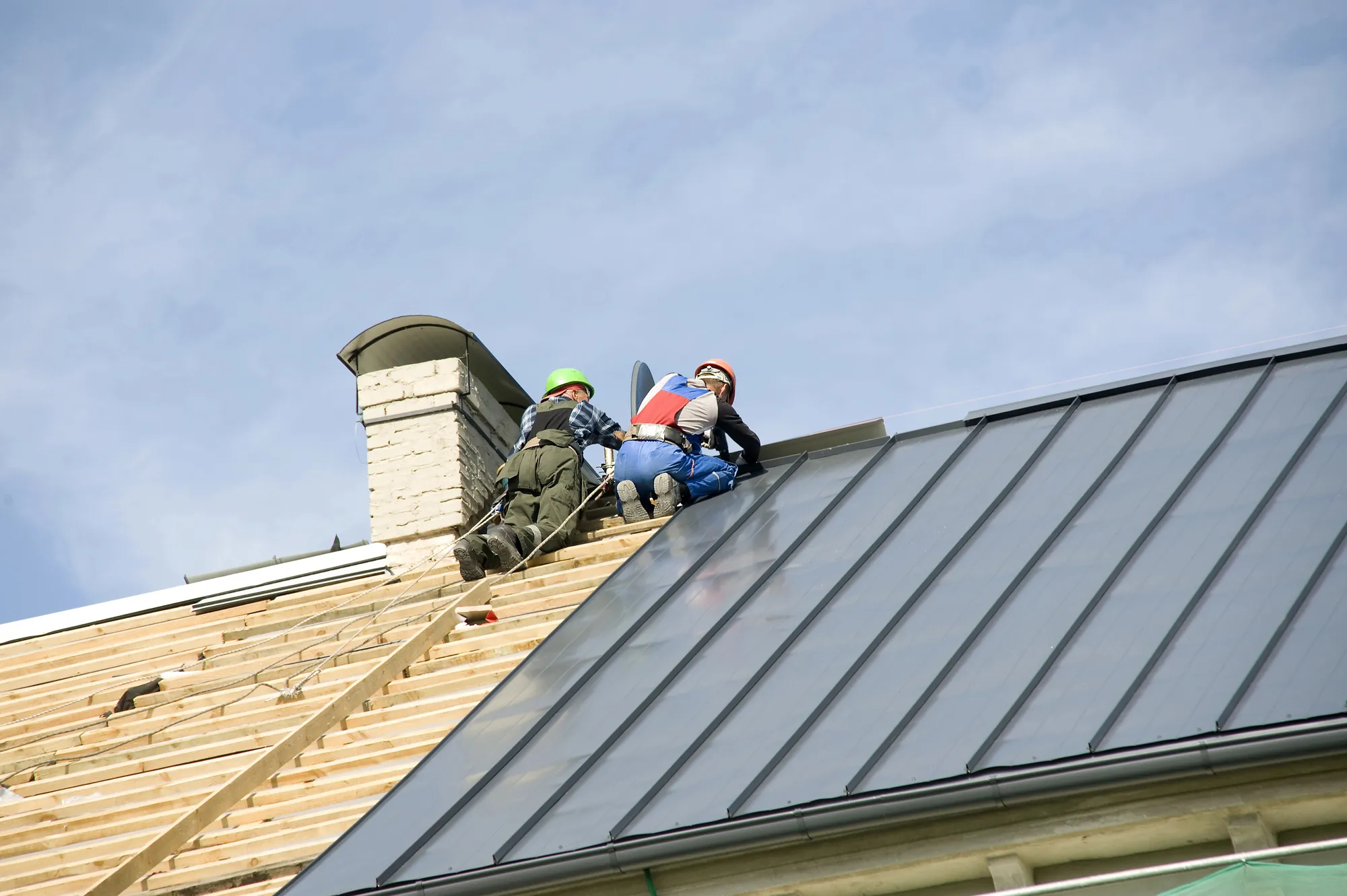 Two roofers wearing helmets and harnesses installing metal roofing panels near a chimney on a house under construction
