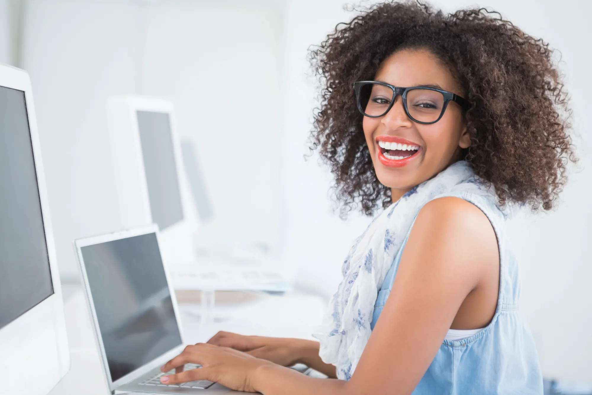 Smiling woman wearing glasses typing on a laptop in an office with computer monitors in the background