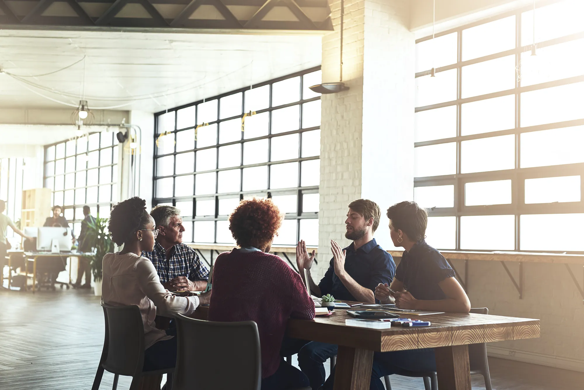 Team of five colleagues having a discussion around a wooden table in a bright, sunlit office