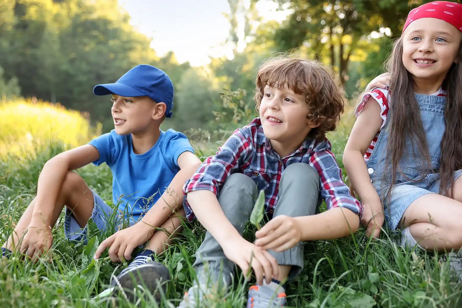 Drei Kinder sitzen entspannt im Gras in einem sonnigen Park oder Waldgebiet.