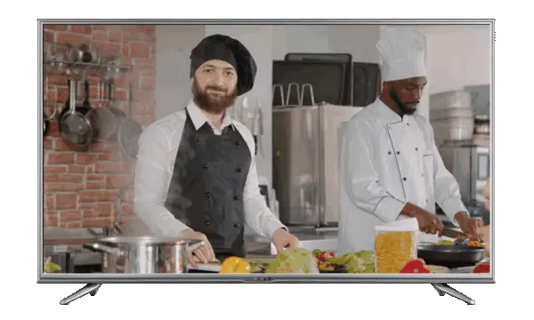 Two male chefs cooking in a kitchen, one smiling at the camera, the other focused on a pan.