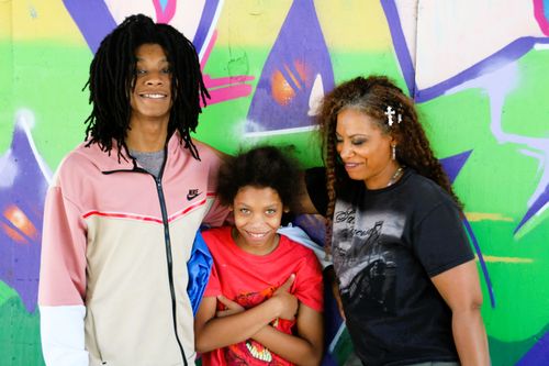 Three people smiling and posing in front of a colorful graffiti wall, including a woman with long braided hair and two children.