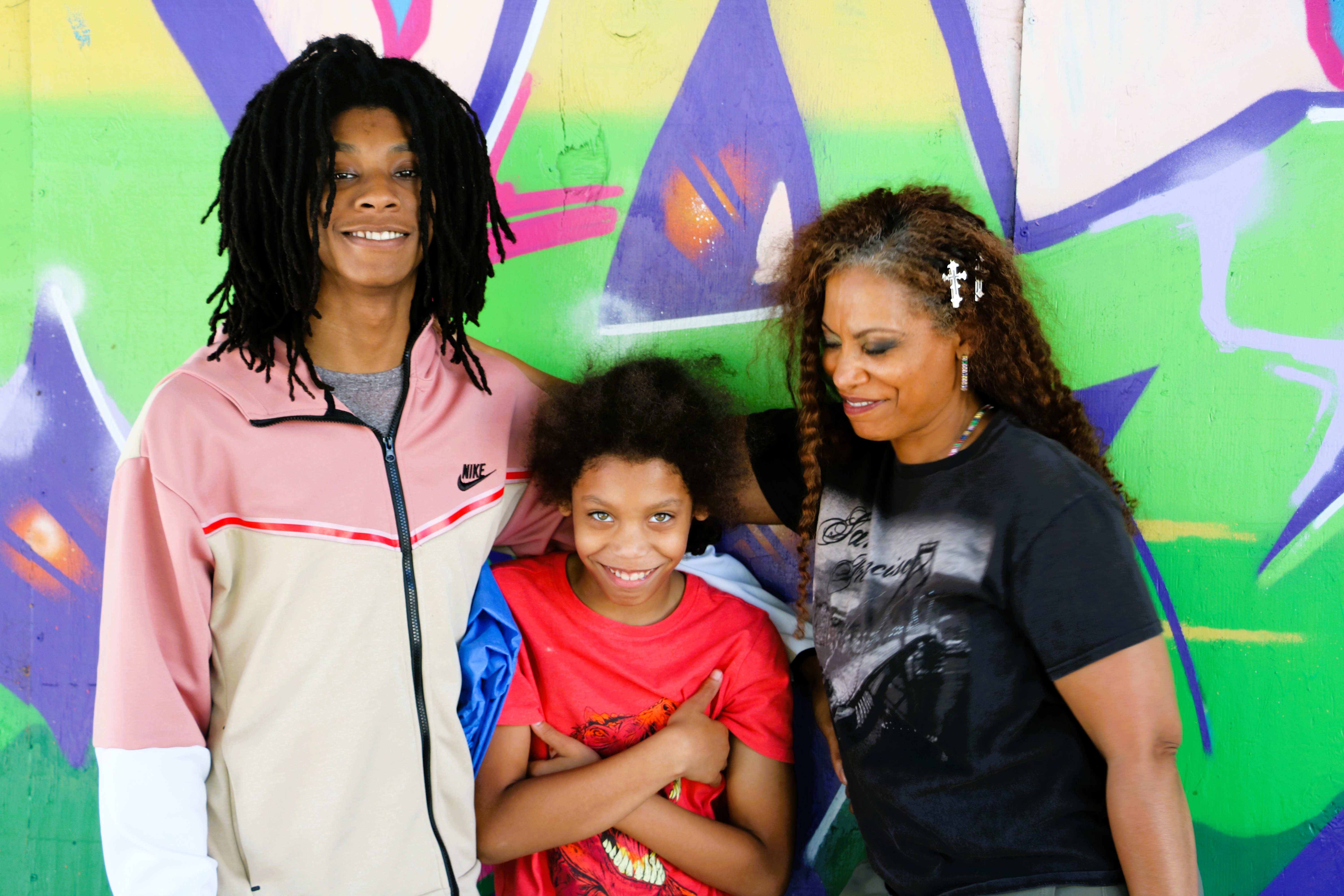 Three people smiling and posing in front of a colorful graffiti wall, including a woman with long braided hair and two children.