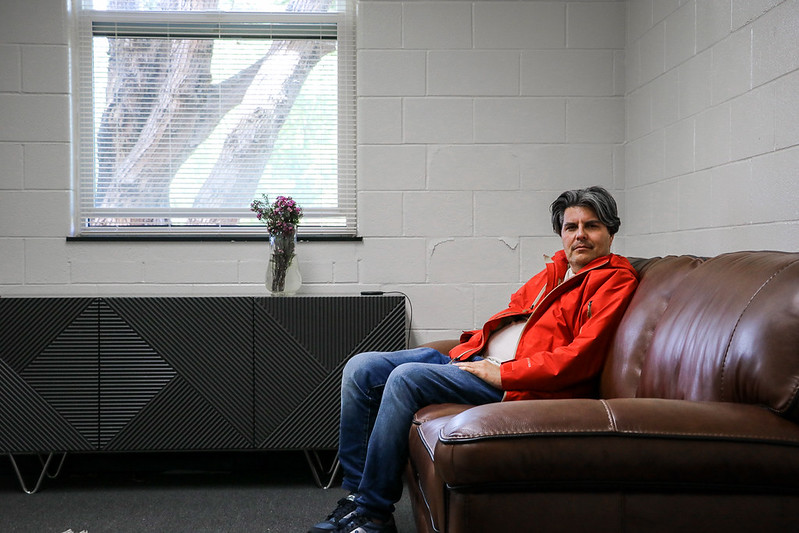 Man in a red jacket sitting on a brown leather couch in a room with a window, a black console table, and a vase with flowers.