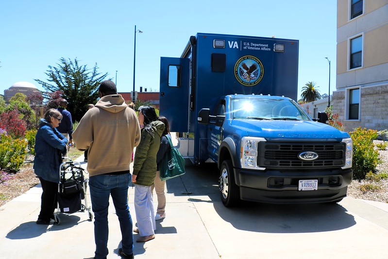 People gathered outside a blue U.S. Department of Veterans Affairs mobile health unit truck in a sunny urban setting.
