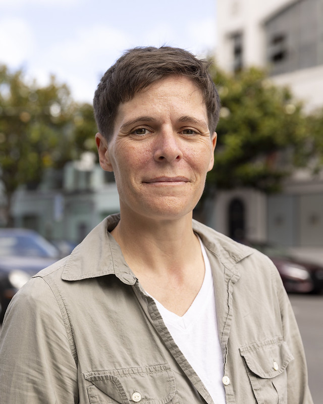 Portrait of a person with short brown hair wearing a light gray shirt and white t-shirt standing outdoors with blurred cars and buildings in the background.