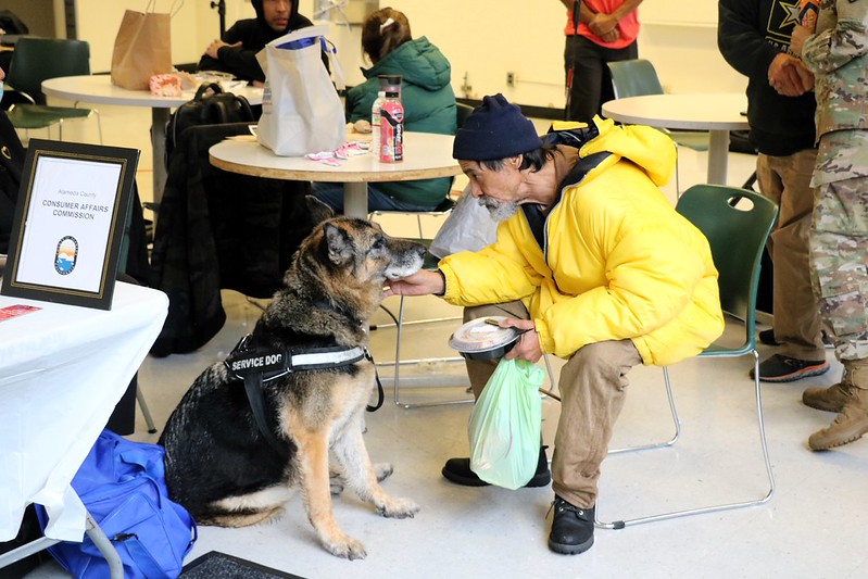 Man in a yellow jacket petting a German shepherd service dog indoors with people seated at tables in the background.