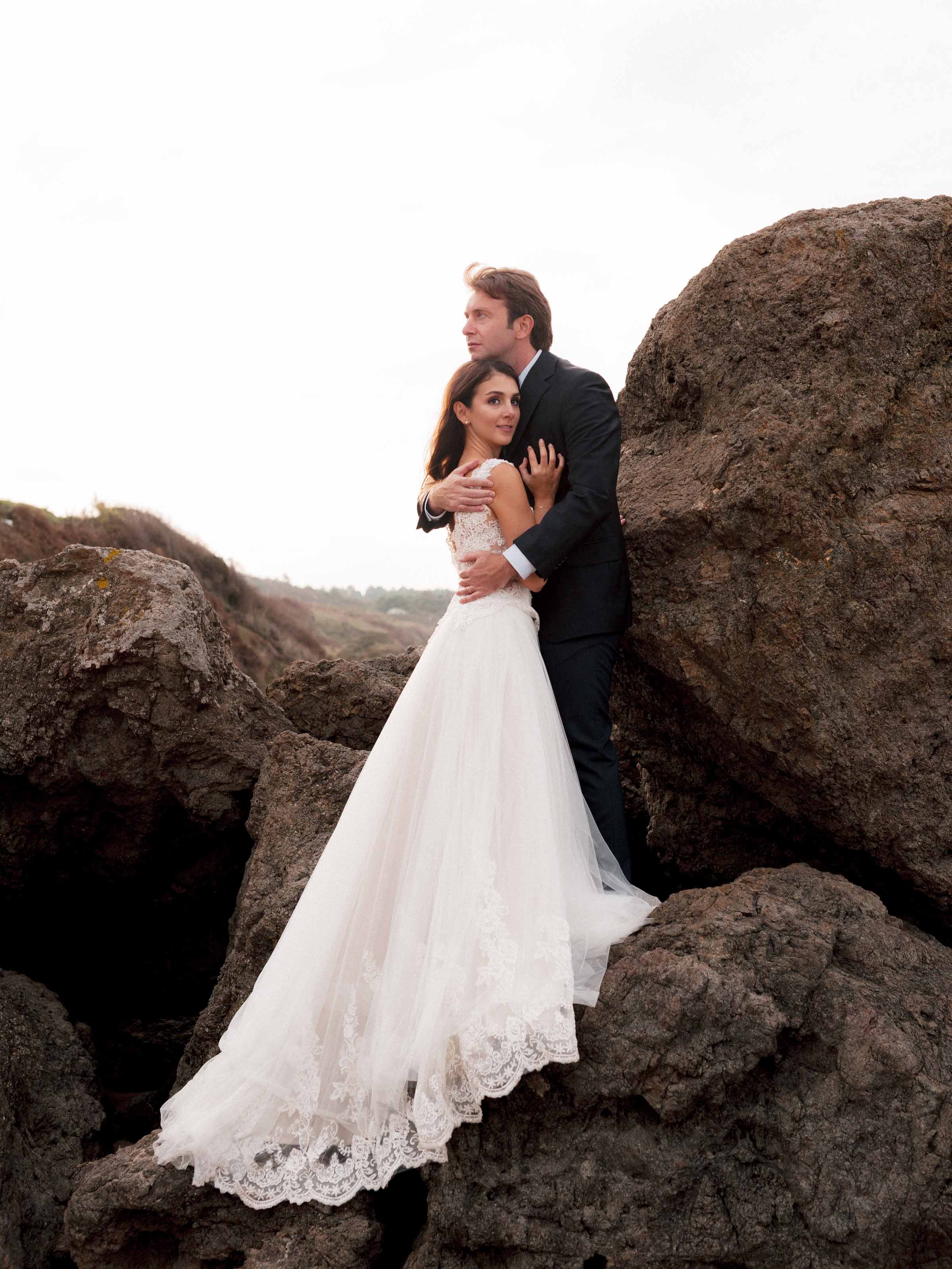 Couple embracing against dark coastal rocks, the bride's lace train spread below, Istanbul