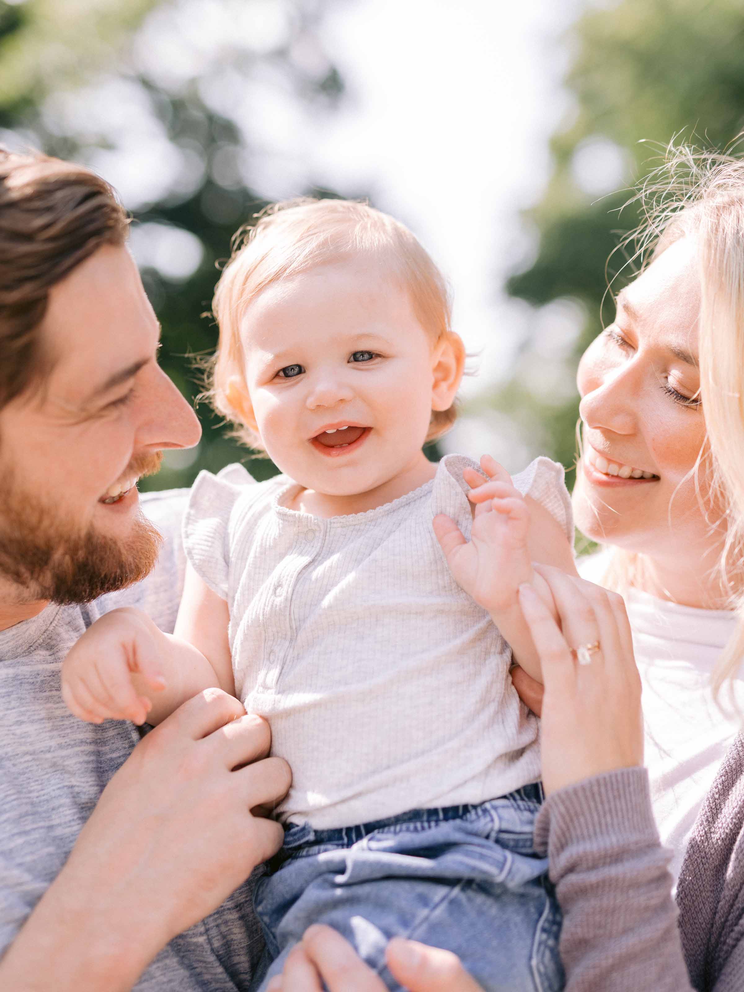 Baby held between both parents, all three laughing under summer trees, Montréal