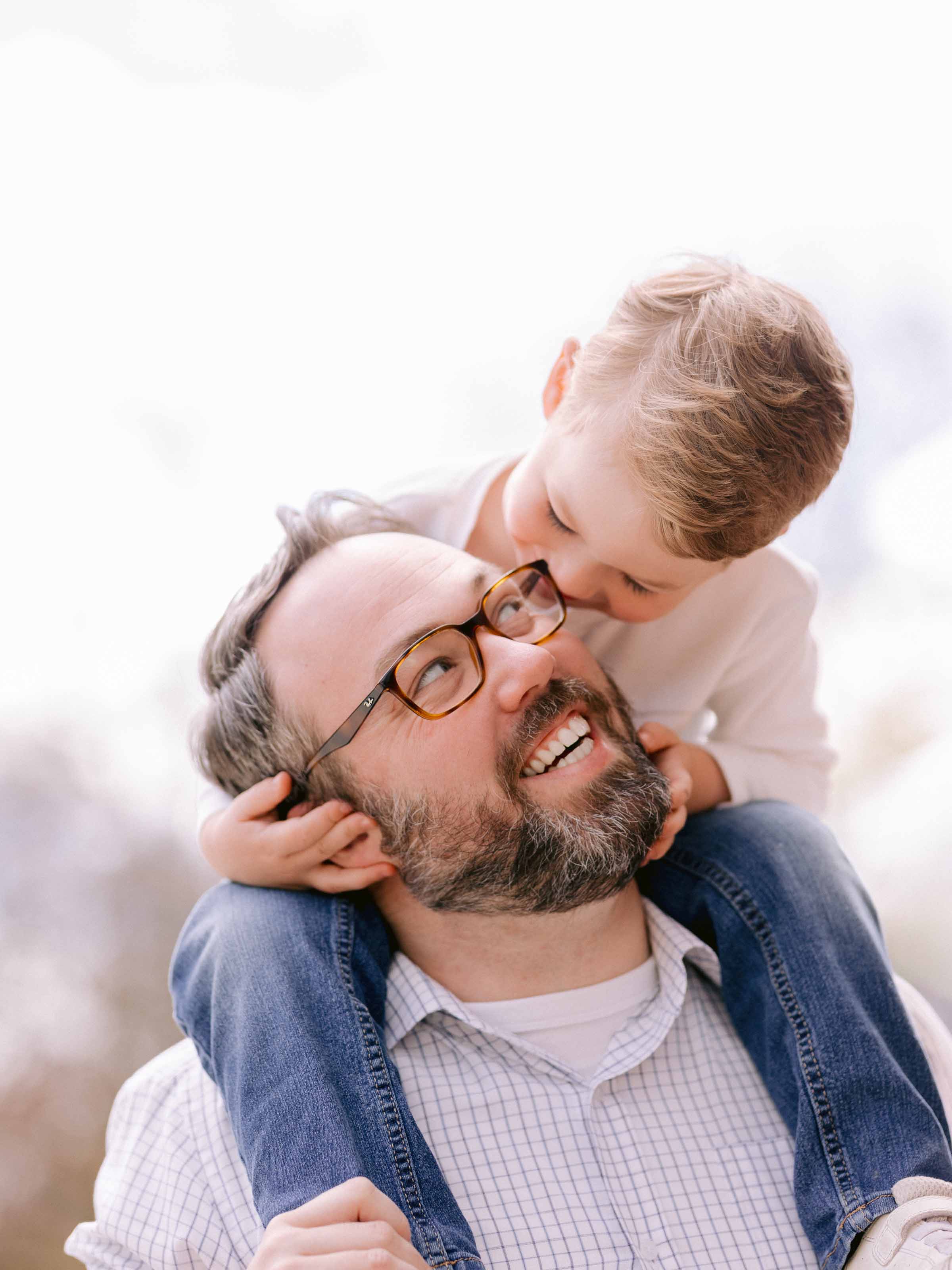 Young boy on his father’s shoulders leaning down to kiss his cheek in bright winter light, Montréal