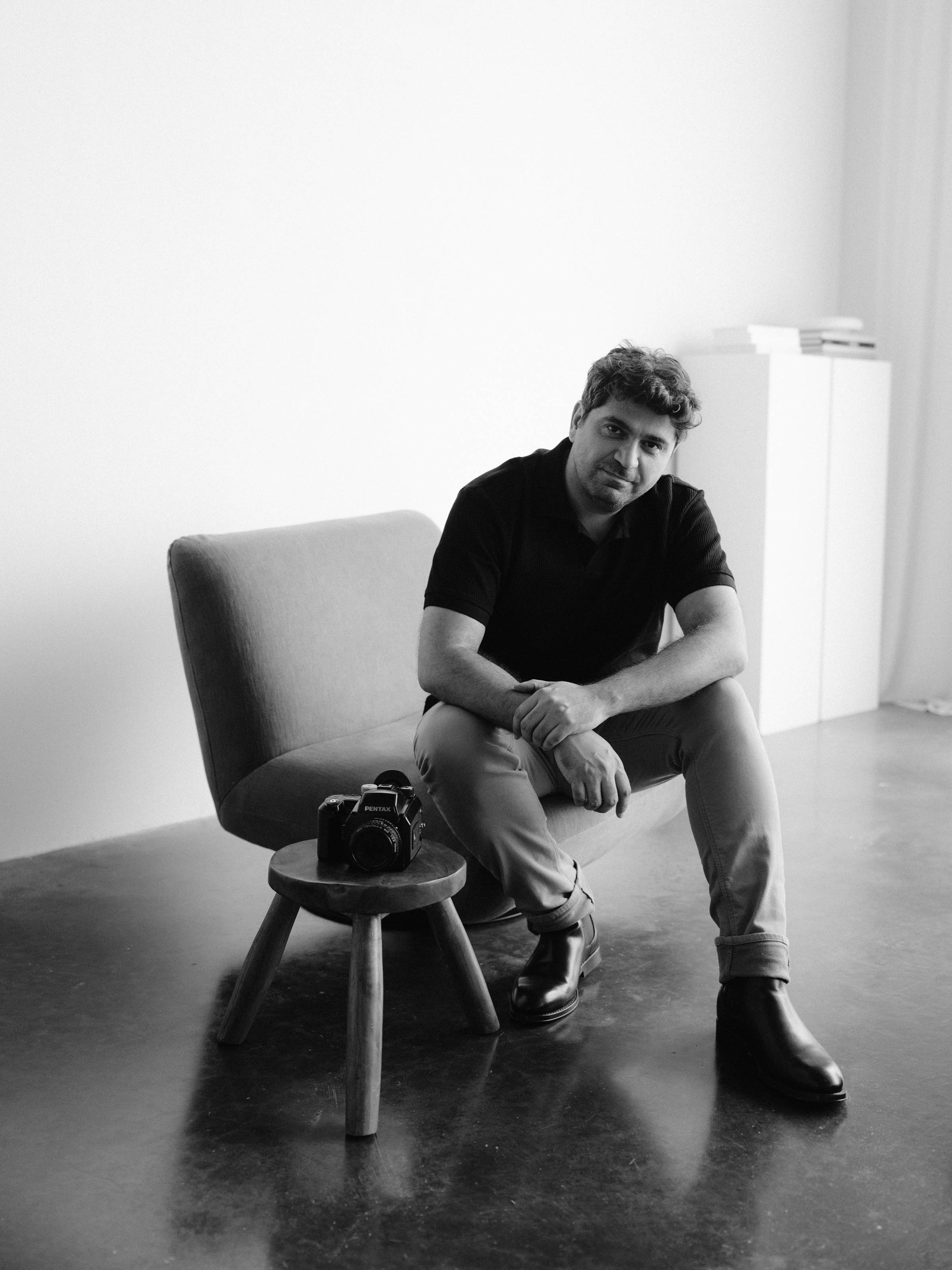 Cagdas Yoldas seated in a bright studio beside a Pentax film camera resting on a wooden stool, black and white