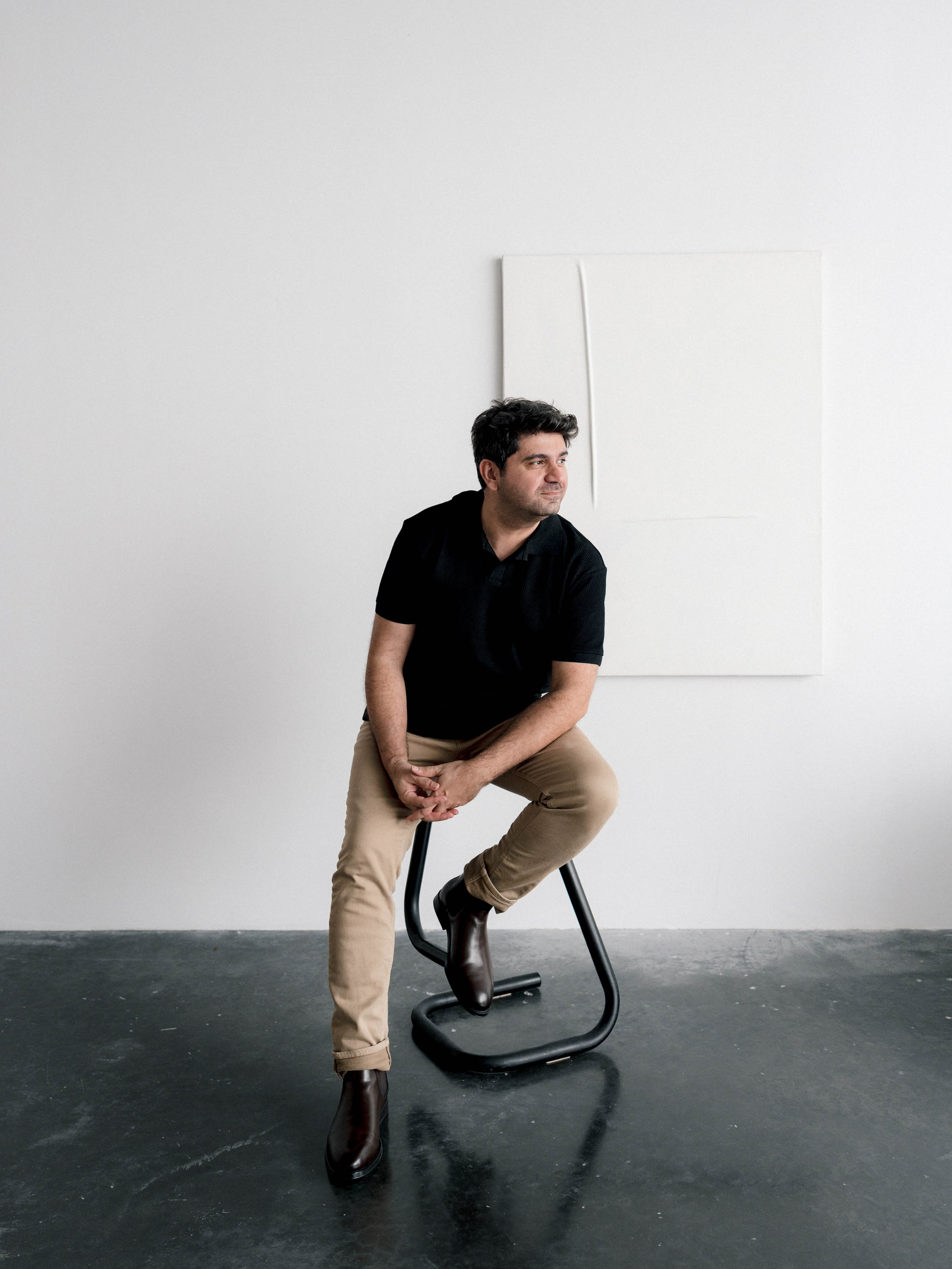 Cagdas Yoldas perched on a stool against a white gallery wall, looking off to one side