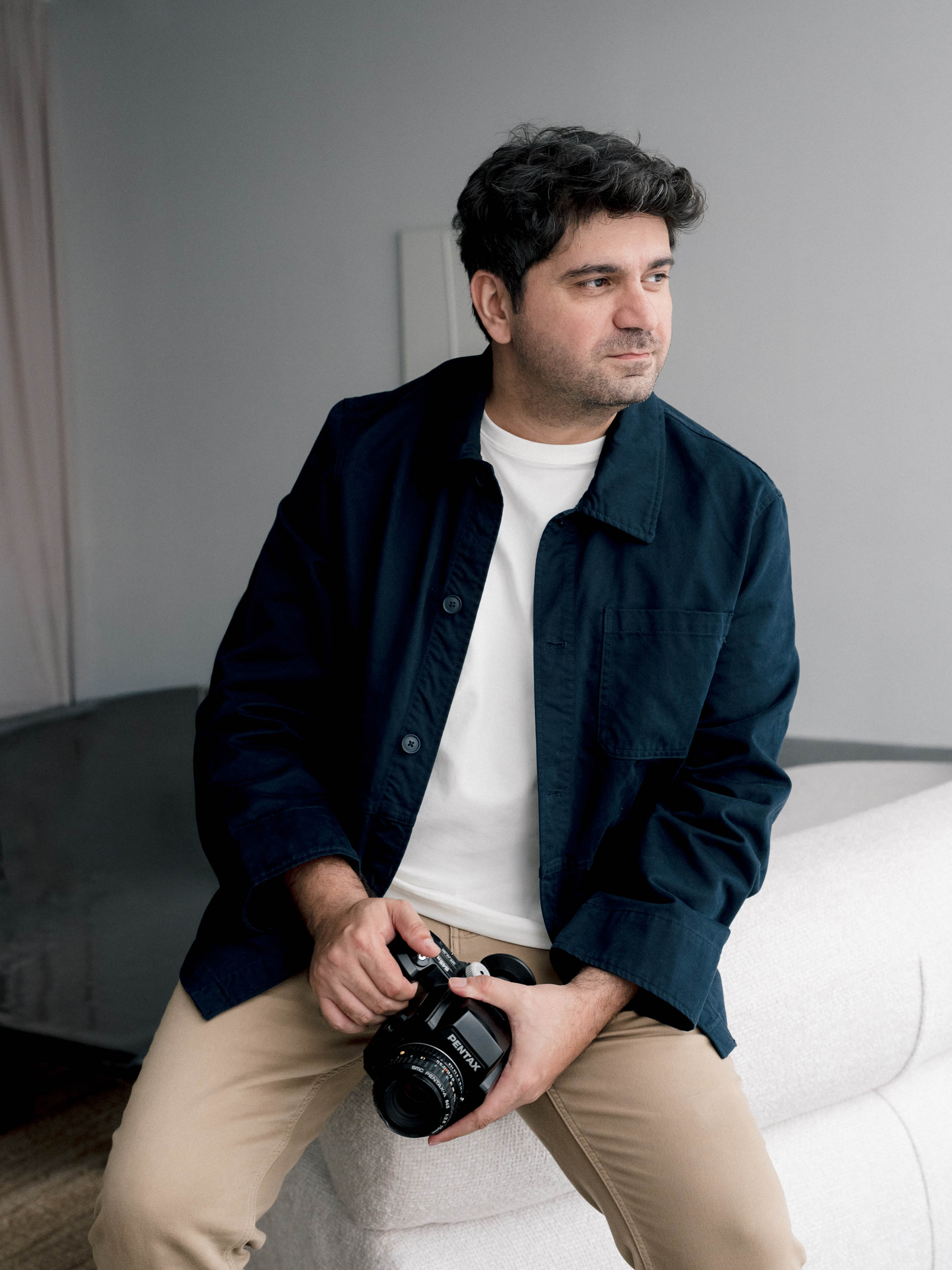 Cagdas Yoldas in a navy jacket, holding a Pentax medium format film camera and looking towards the window light