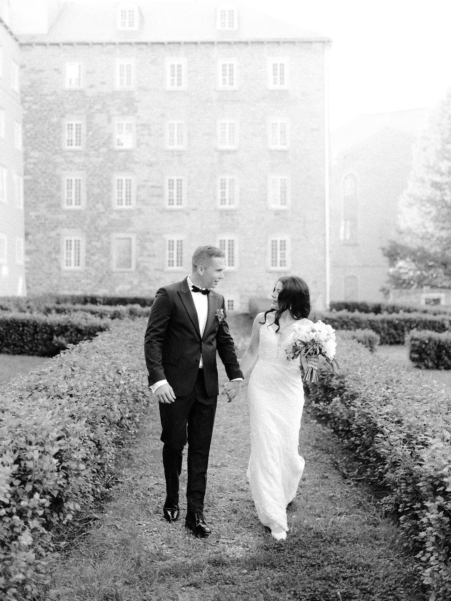 Bride and groom walking hand in hand between clipped hedges beside the stone seminary in Old Quebec