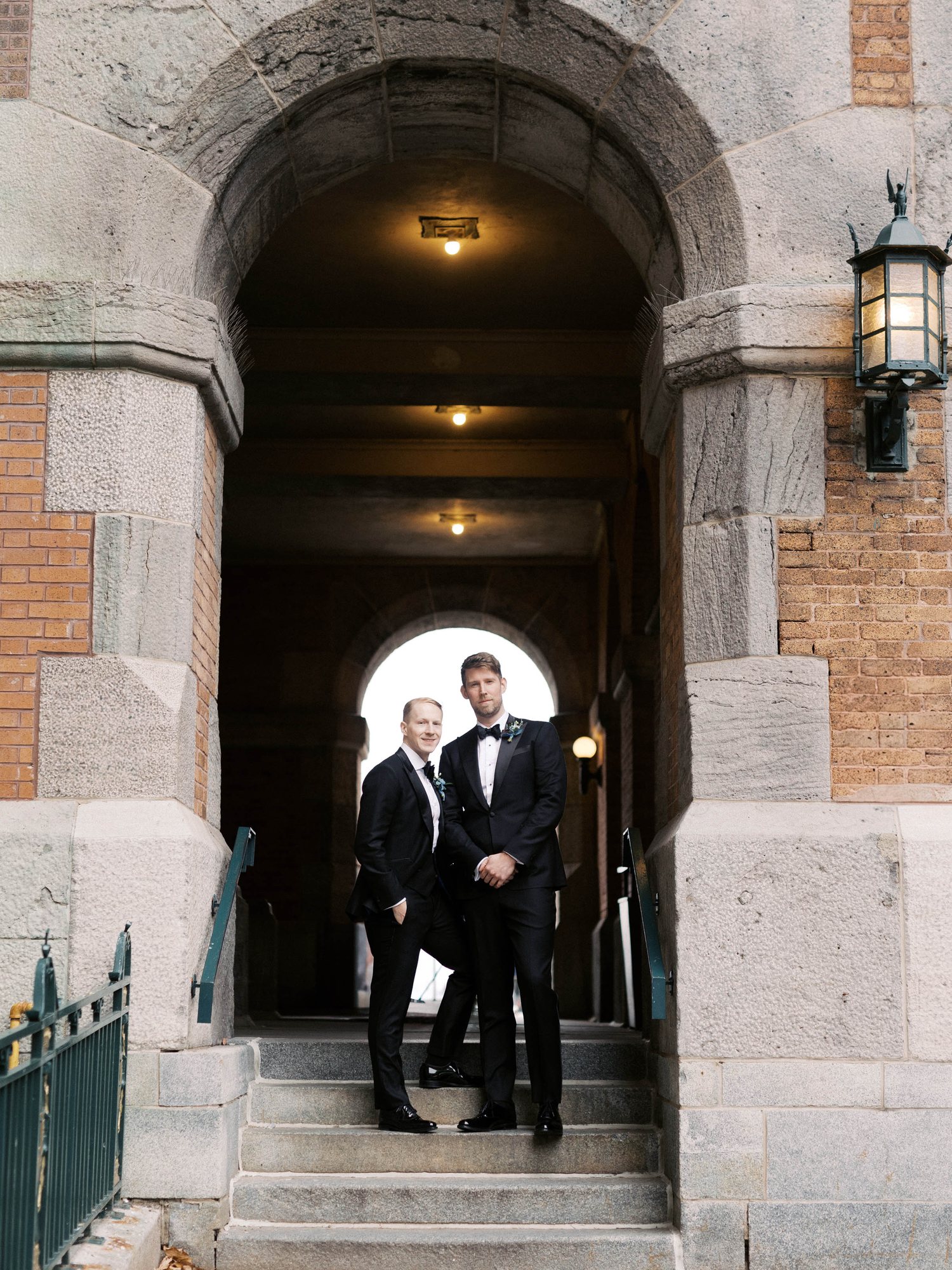 Two grooms standing together in a stone archway lit by an iron lantern in Old Quebec