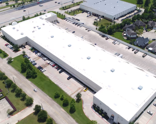 Aerial view of a large white industrial warehouse with surrounding parking lots, nearby roads, and adjacent smaller buildings.