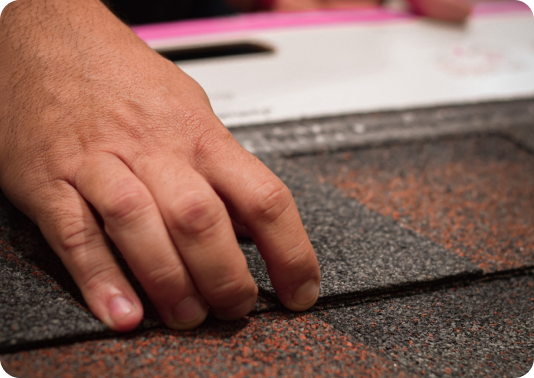 A hand lifting and inspecting dark asphalt roofing shingles on a roof.