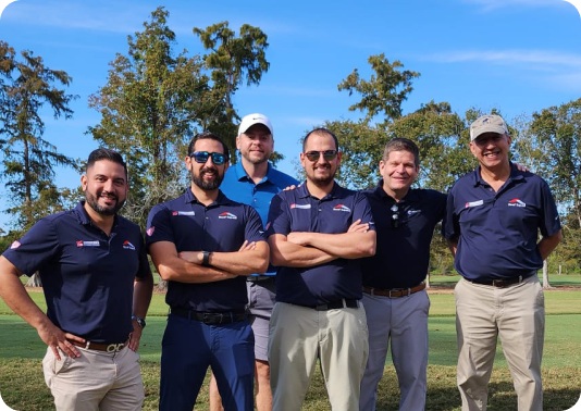 Group of six men wearing navy polo shirts standing outdoors on a grassy area with trees and blue sky in the background.