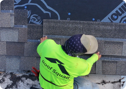 Worker wearing a bright green Roof Squad shirt installing asphalt shingles on a roof.