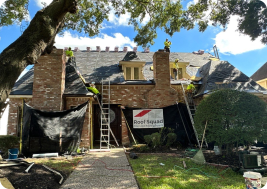 Workers installing a new roof on a brick house under a partly cloudy sky, with ladders and protective tarps in place.